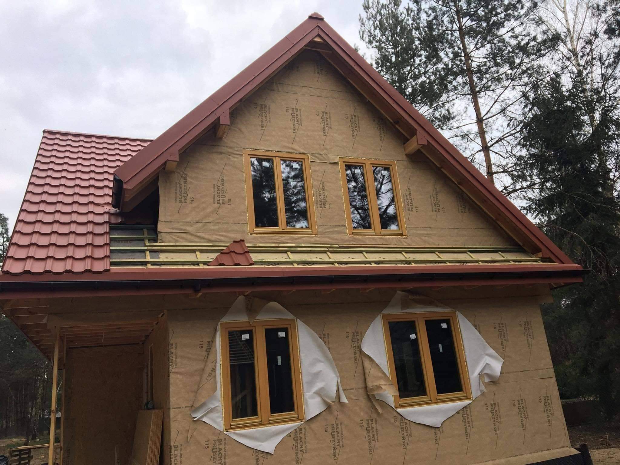 Dom w trakcie budowy z częściowo położonym dachem z czerwonej dachówki, zabezpieczonymi oknami i membraną dachową na ścianach.