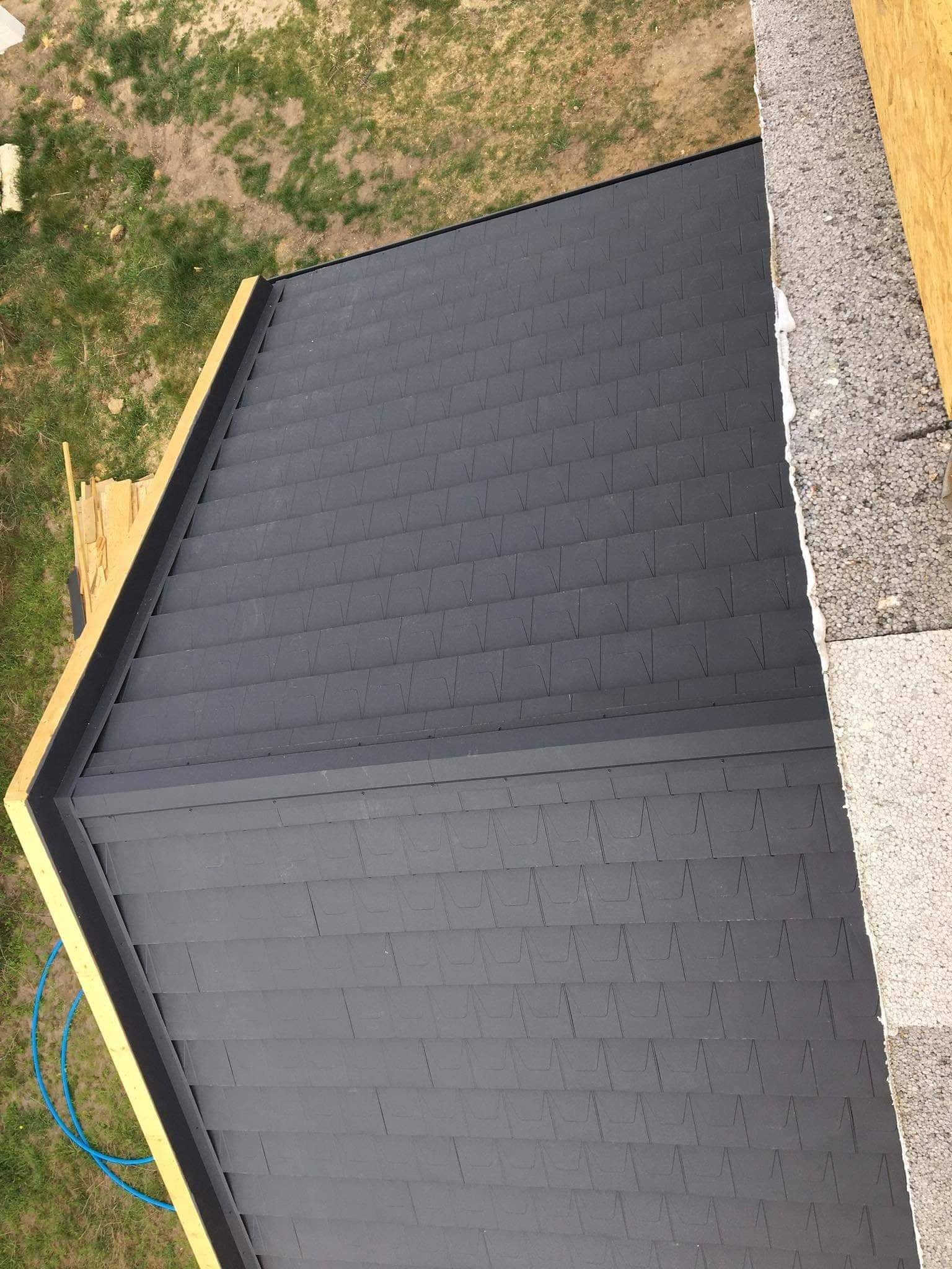 Widok z góry na fragment dachu pokrytego ciemnoszarą dachówką bitumiczną, widoczne obróbki blacharskie i fragment ściany budynku z izolacją.