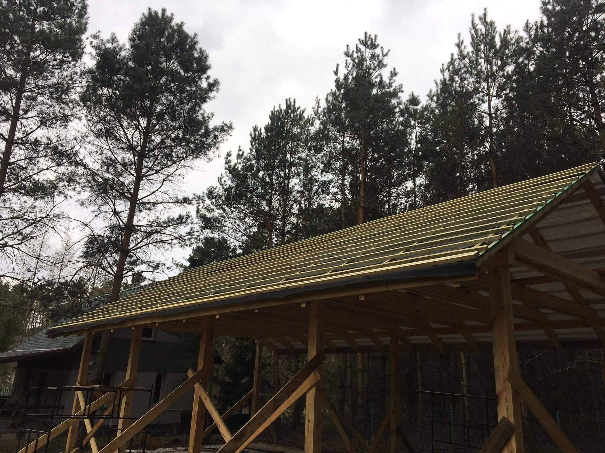 Konstrukcja drewniana dachu w trakcie budowy z widocznymi łatami i membraną, otoczona lasem iglastym przy pochmurnym niebie.