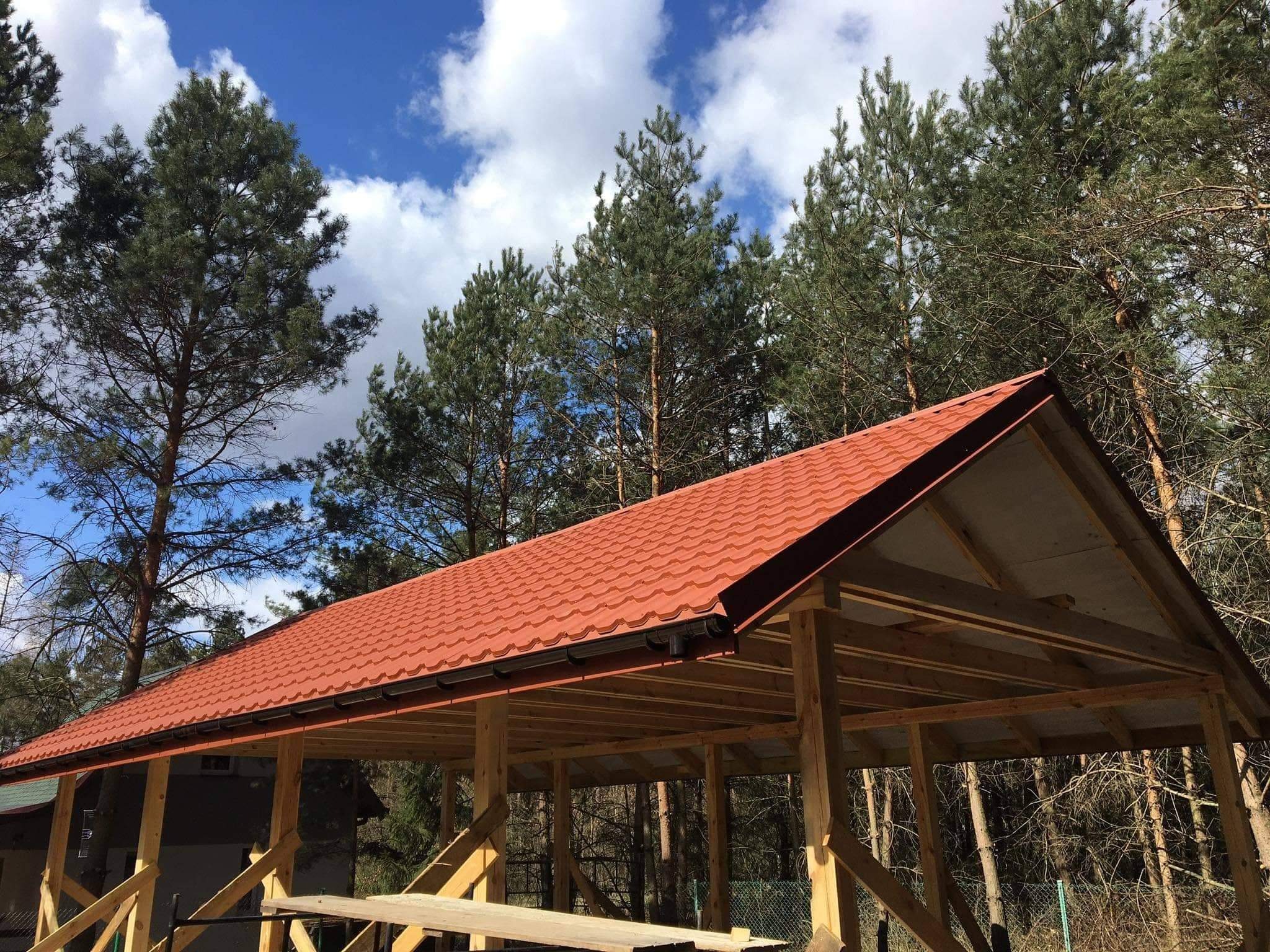 Czerwony dach z blachy trapezowej, z zamontowanymi rynnami, na drewnianej konstrukcji w otoczeniu drzew iglastych i błękitnego nieba z chmurami.