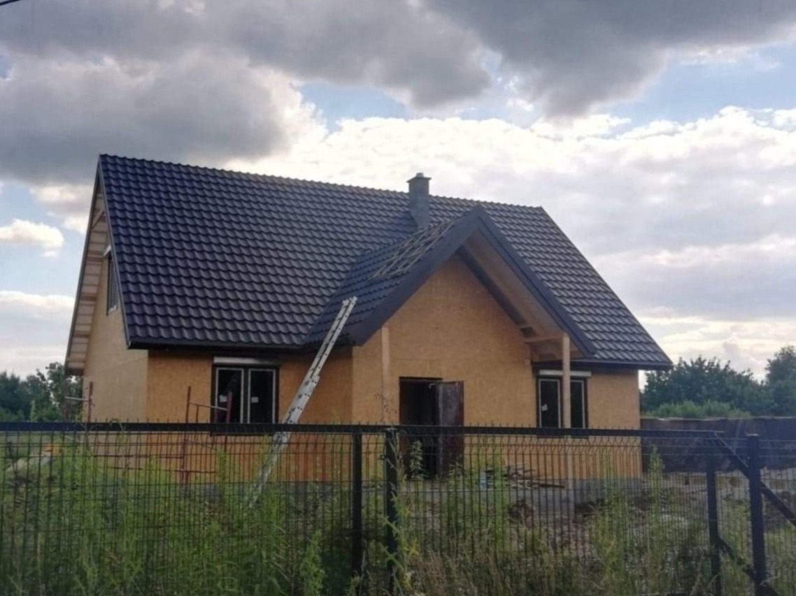 Dom w trakcie budowy z nowym, ciemnym dachem z blachodachówki, drabina oparta o dach, widoczne surowe ściany z płyt OSB, ogrodzenie z siatki na pierwszym planie.