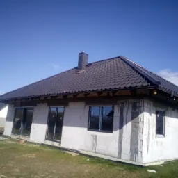 Niedokończony dom murowany z ciemną dachówką i zamontowanymi oknami, widok z zewnątrz na tle błękitnego nieba.