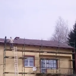 Prace dekarskie na dachu domu jednorodzinnego z użyciem rusztowania i drabiny, widoczni dwaj dekarze w trakcie układania brązowej dachówki.