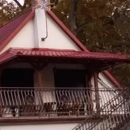 Dwupiętrowy dom z czerwonym dachem, balkonem z metalową balustradą i ceglanym kominem, widok z dołu na tle drzew.