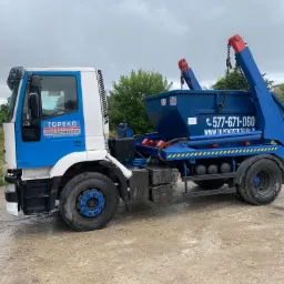 Ciężarówka z niebieskim kontenerem na platformie, z logo firmy TOPEKO STRZELIN, zaparkowana na betonowym placu w pochmurny dzień.