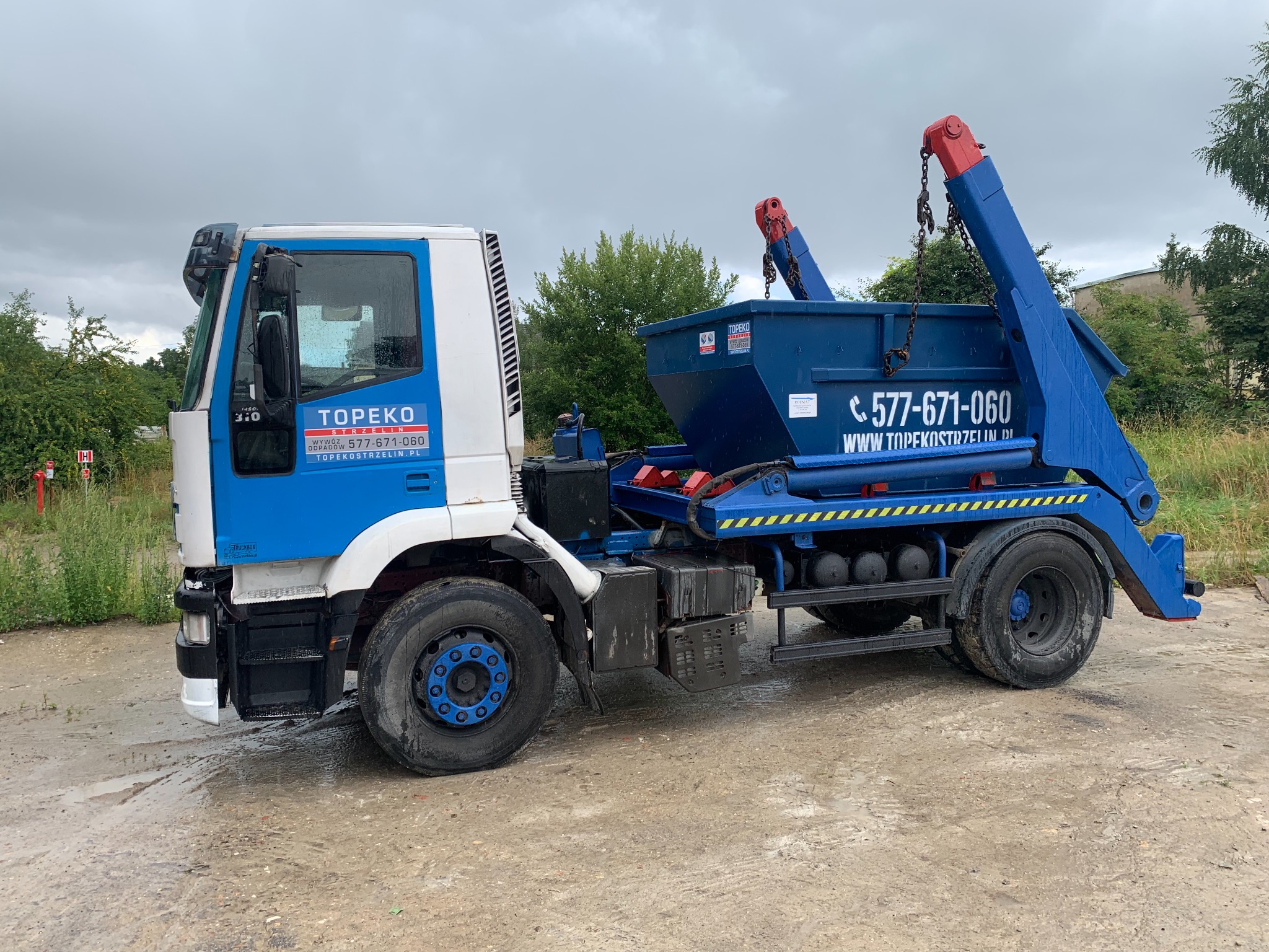 Ciężarówka z niebieskim kontenerem na platformie, z logo firmy TOPEKO STRZELIN, zaparkowana na betonowym placu w pochmurny dzień.