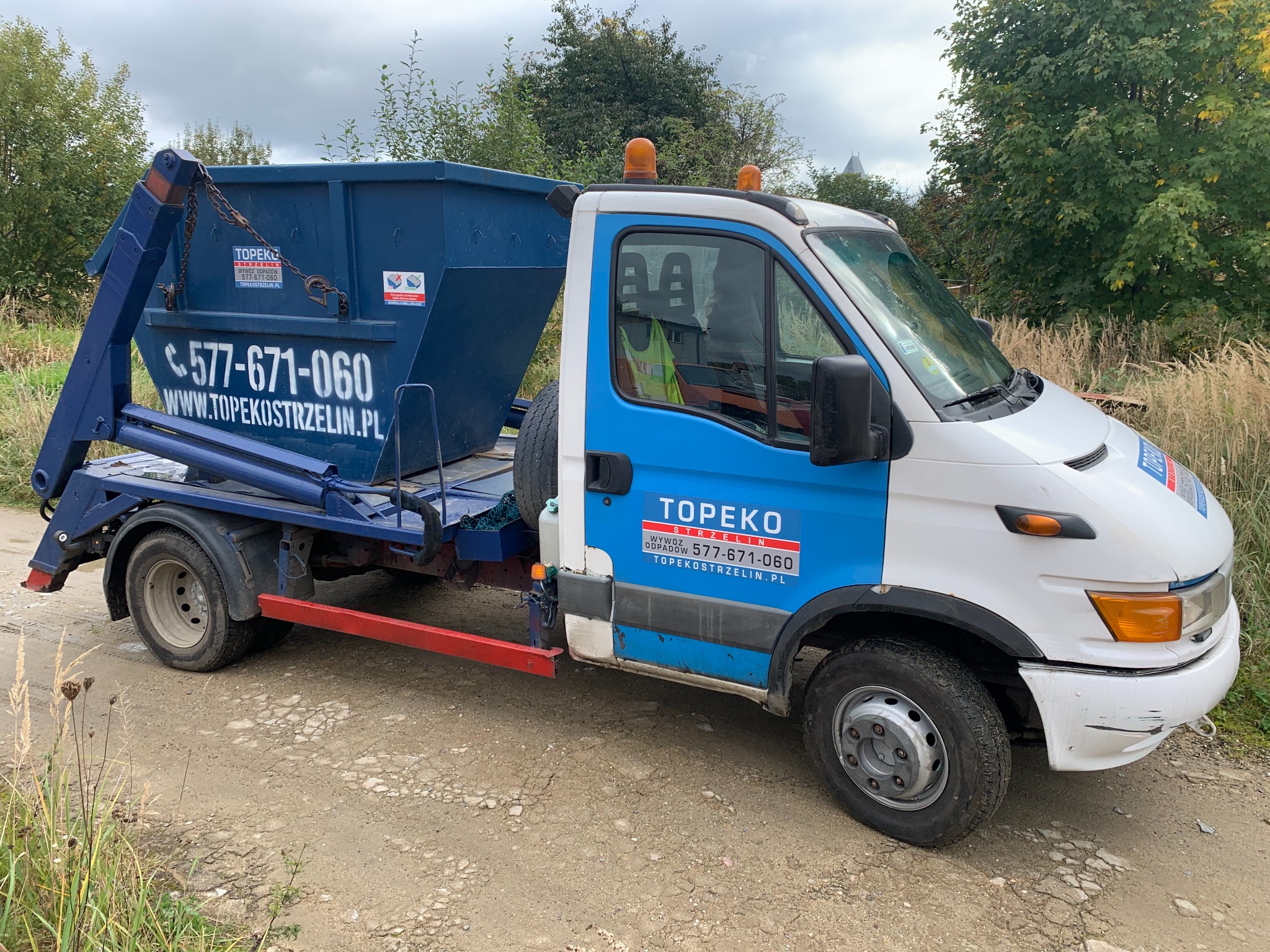 Ciężarówka z niebieskim kontenerem na gruz, z widocznym numerem telefonu i adresem strony internetowej firmy Topekostrzelin.pl, stojąca na polnej drodze.