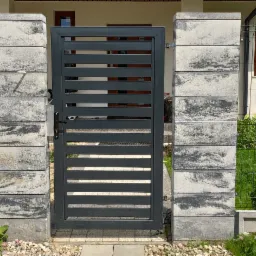 Nowoczesna, szara brama z poziomymi szczeblami, osadzona między dwoma słupkami z szarej kostki imitującej kamień, z widocznym adresem na ogrodzeniu z siatki w tle.