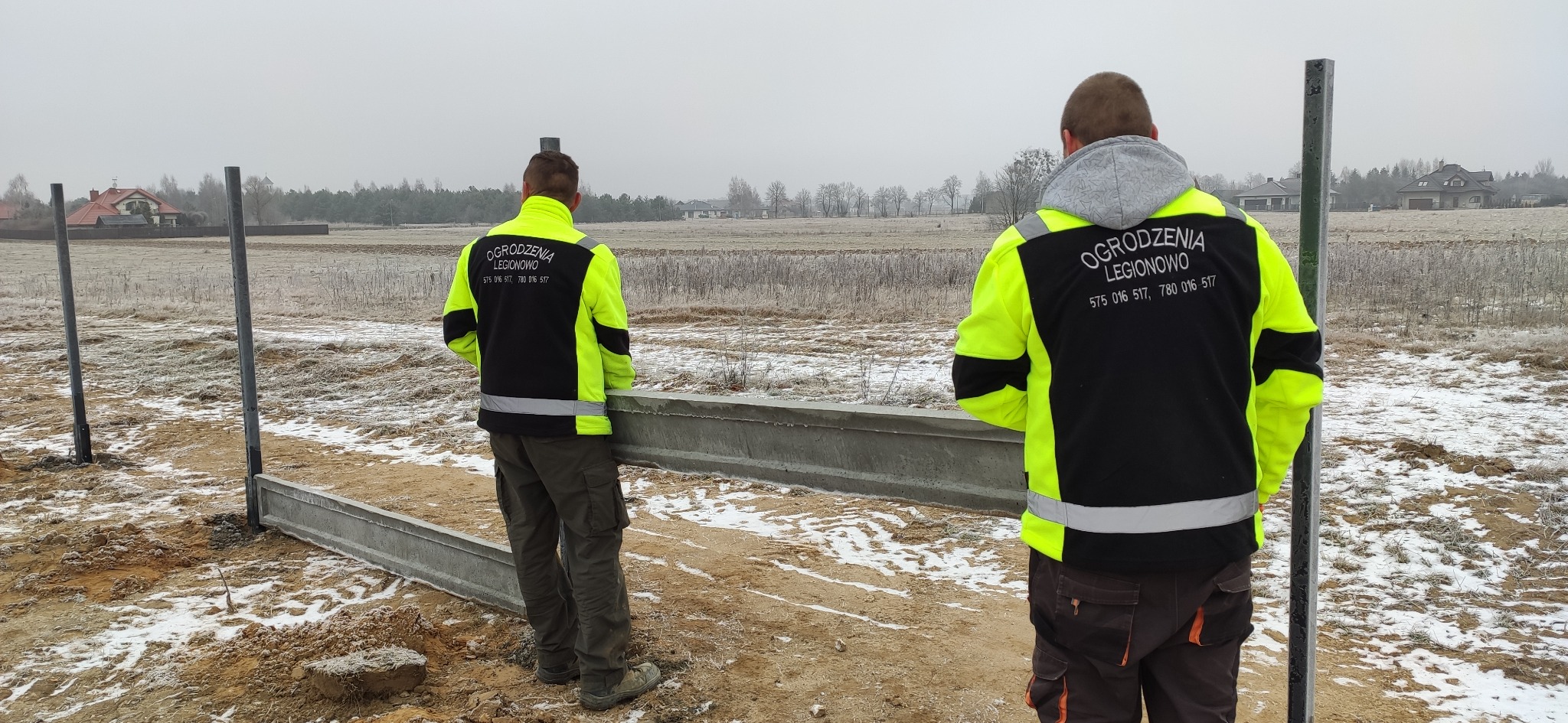 Dwóch pracowników w odblaskowych kurtkach z logo firmy 'Ogrodzenia Legionowo' montuje betonową podmurówkę ogrodzenia na polu pokrytym resztkami śniegu.