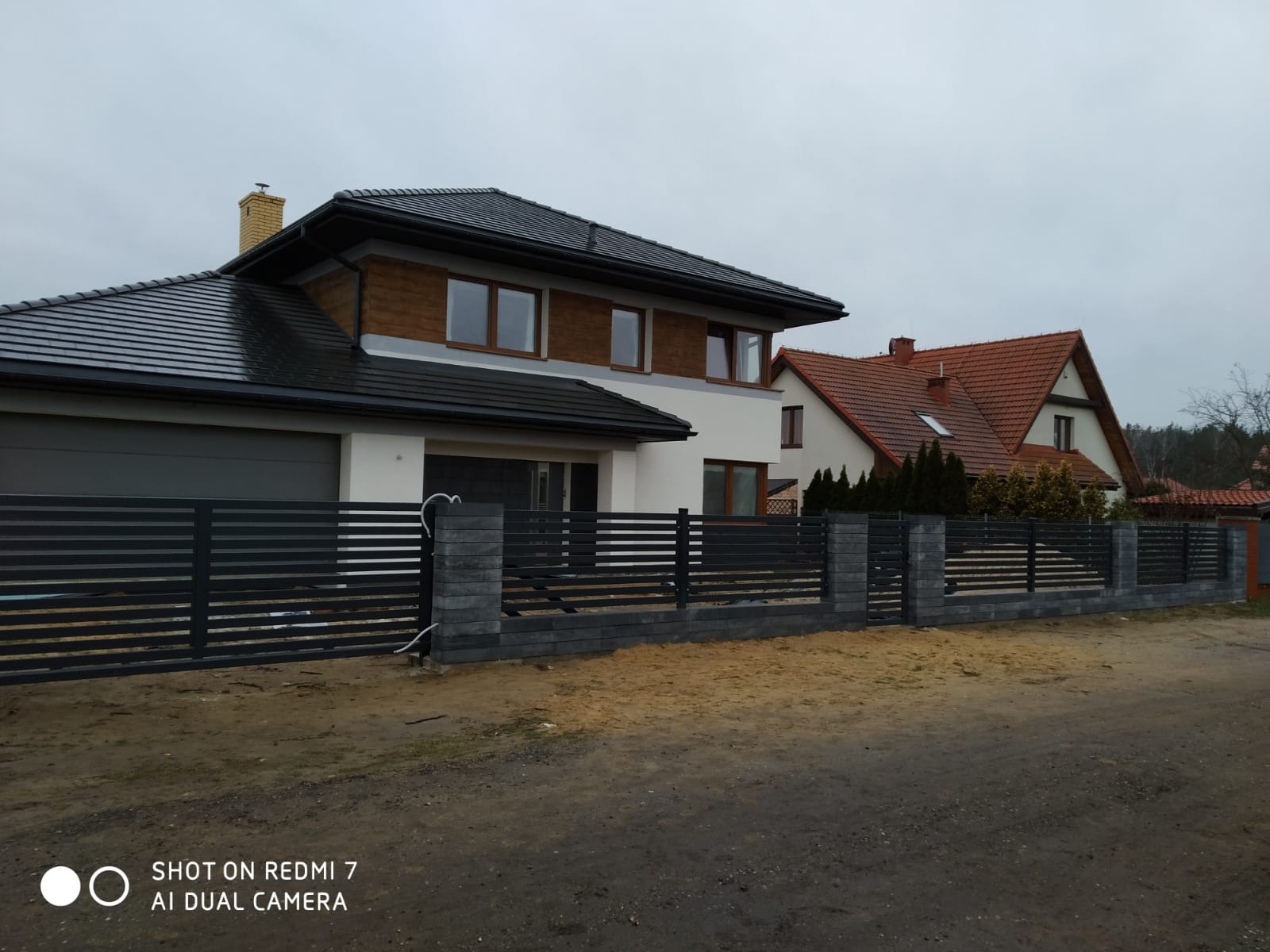 Nowoczesne ogrodzenie panelowe w kolorze antracytowym z betonowymi słupkami, widoczne na tle domu z garażem i sąsiedniego domu z czerwonym dachem, sfotografowane w pochmurny dzień.