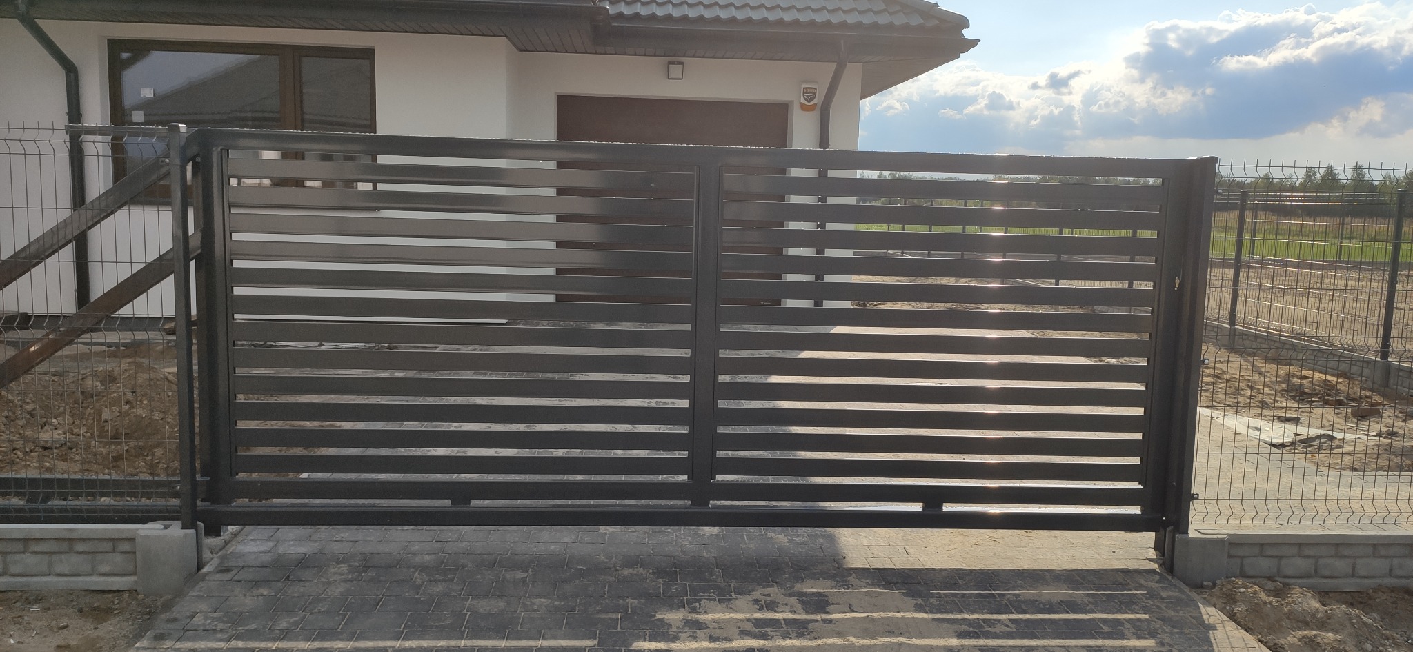 Nowoczesna, przesuwna brama w kolorze antracytowym z poziomymi panelami, zamontowana na posesji z kostką brukową i ogrodzeniem z siatki.