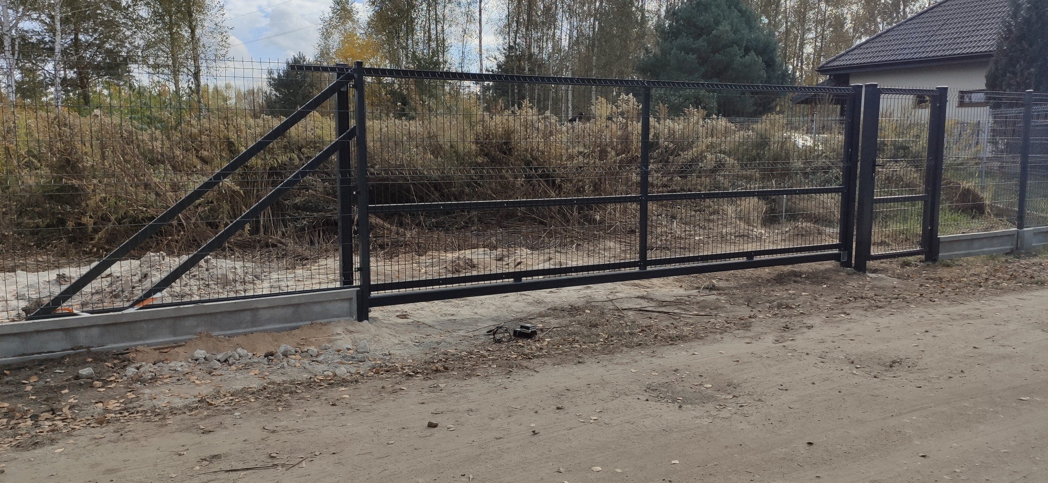 Nowo zamontowana, przesuwna brama panelowa w kolorze grafitowym, z widocznymi elementami betonowej podstawy i otaczającym terenem w trakcie prac wykończeniowych.