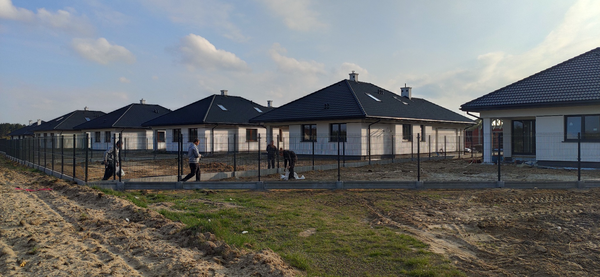 Nowoczesne osiedle domów jednorodzinnych z czarnymi dachami, ogrodzone siatką panelową na betonowej podmurówce, widoczni pracownicy podczas montażu ogrodzenia.