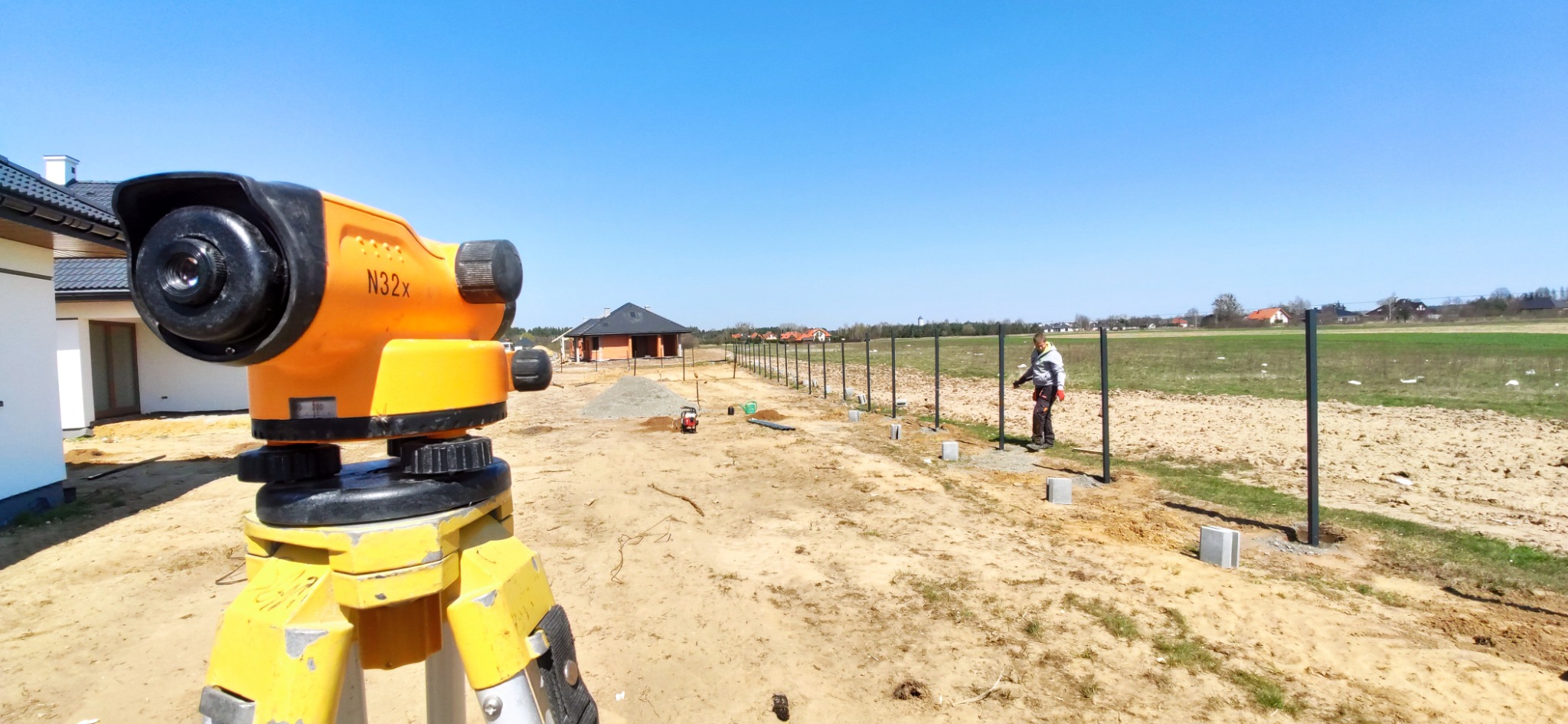 Prace geodezyjne podczas montażu ogrodzenia: niwelator optyczny N32x na statywie w pierwszym planie, w tle pracownik montuje słupki ogrodzeniowe na betonowych podstawach, widok na otwarty teren...