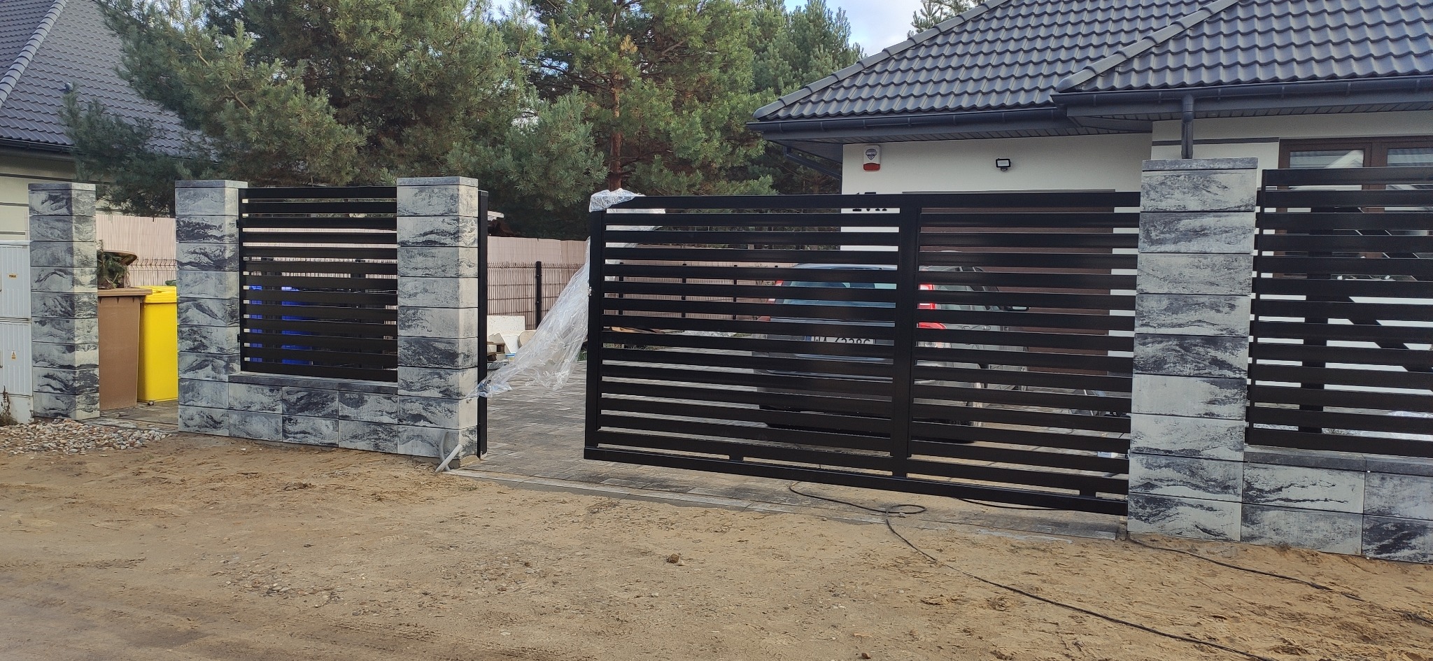 Nowoczesna brama wjazdowa i fragment ogrodzenia panelowego z elementami betonowymi w szaro-czarnej kolorystyce, widoczne ślady montażu w postaci folii ochronnej.