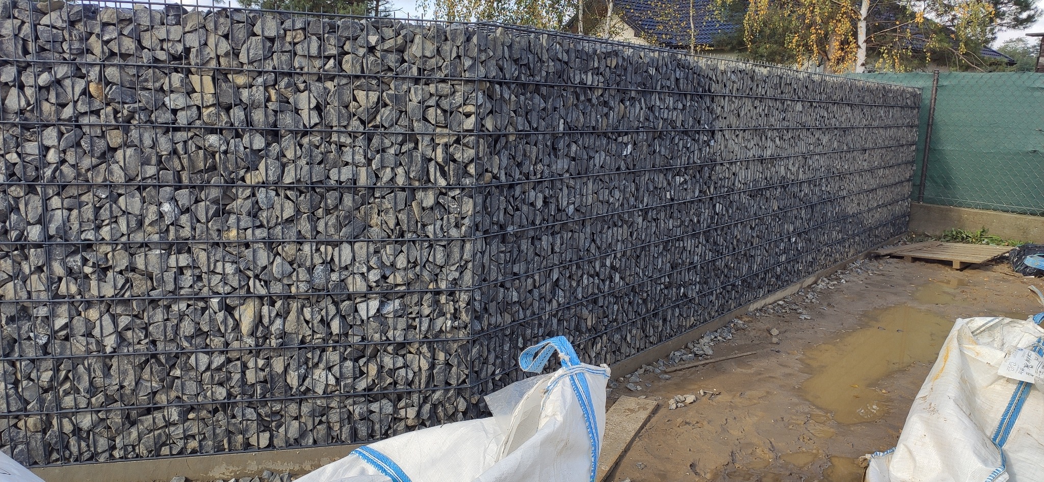 Gabionowe ogrodzenie z szarego kamienia, widoczne worki z materiałem budowlanym i błotniste podłoże.