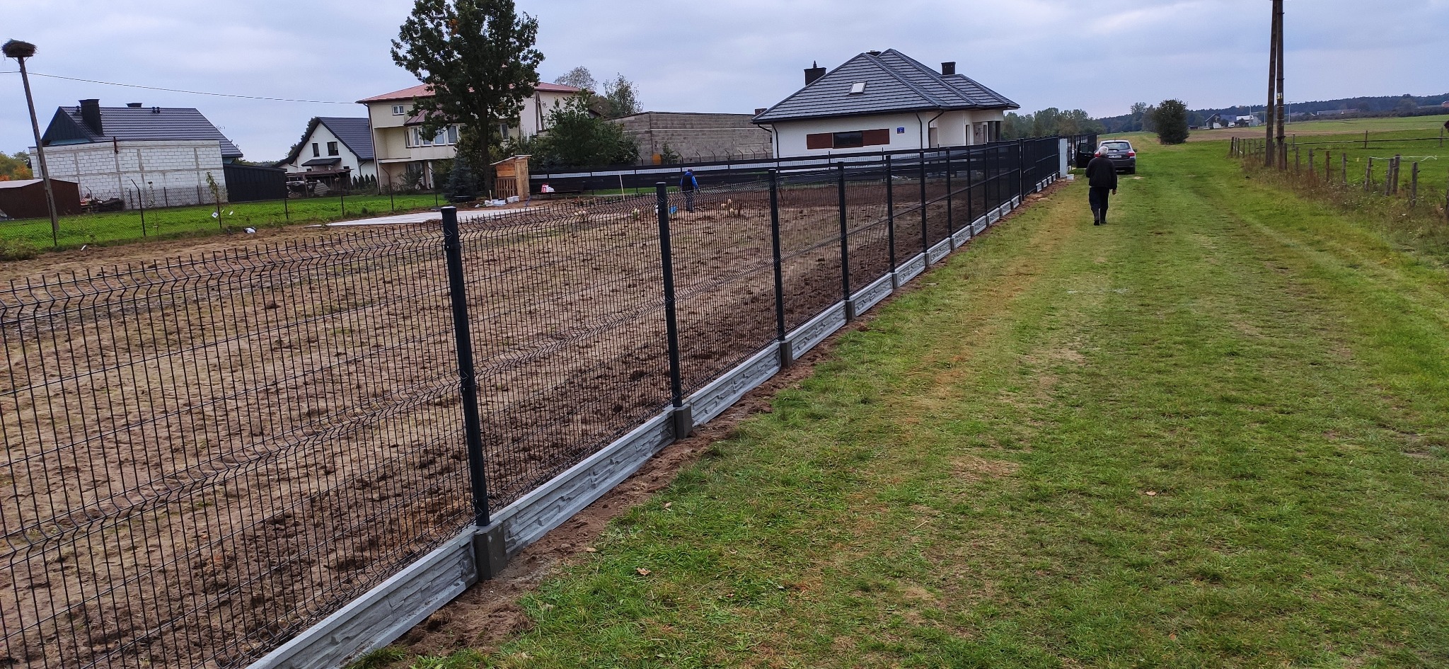 Nowoczesne ogrodzenie panelowe z podmurówką betonową, widok z boku na tle wiejskiego krajobrazu i pochmurnego nieba.