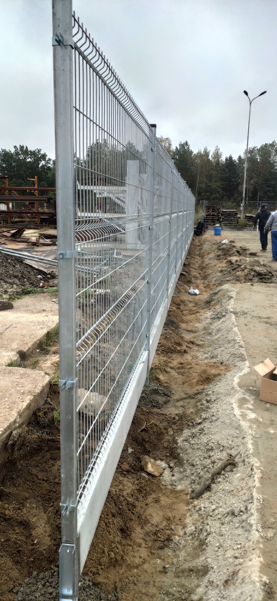 Nowo zainstalowane, metalowe ogrodzenie panelowe z podmurówką betonową, widoczne ślady prac ziemnych wzdłuż linii ogrodzenia.