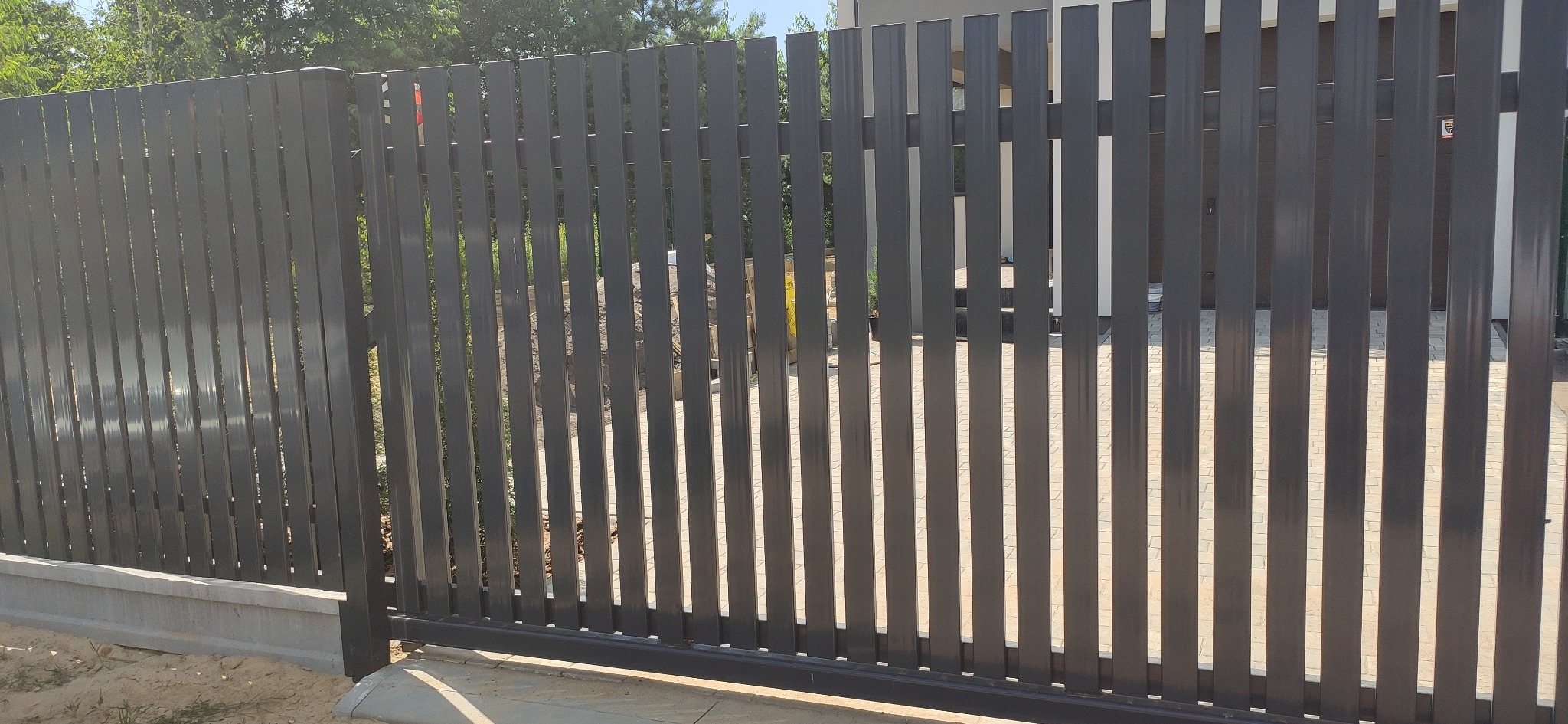 Nowoczesna, przesuwna brama ogrodzeniowa z pionowych, grafitowych profili, zamontowana na betonowej podmurówce, z widocznymi elementami konstrukcyjnymi i otoczeniem posesji.