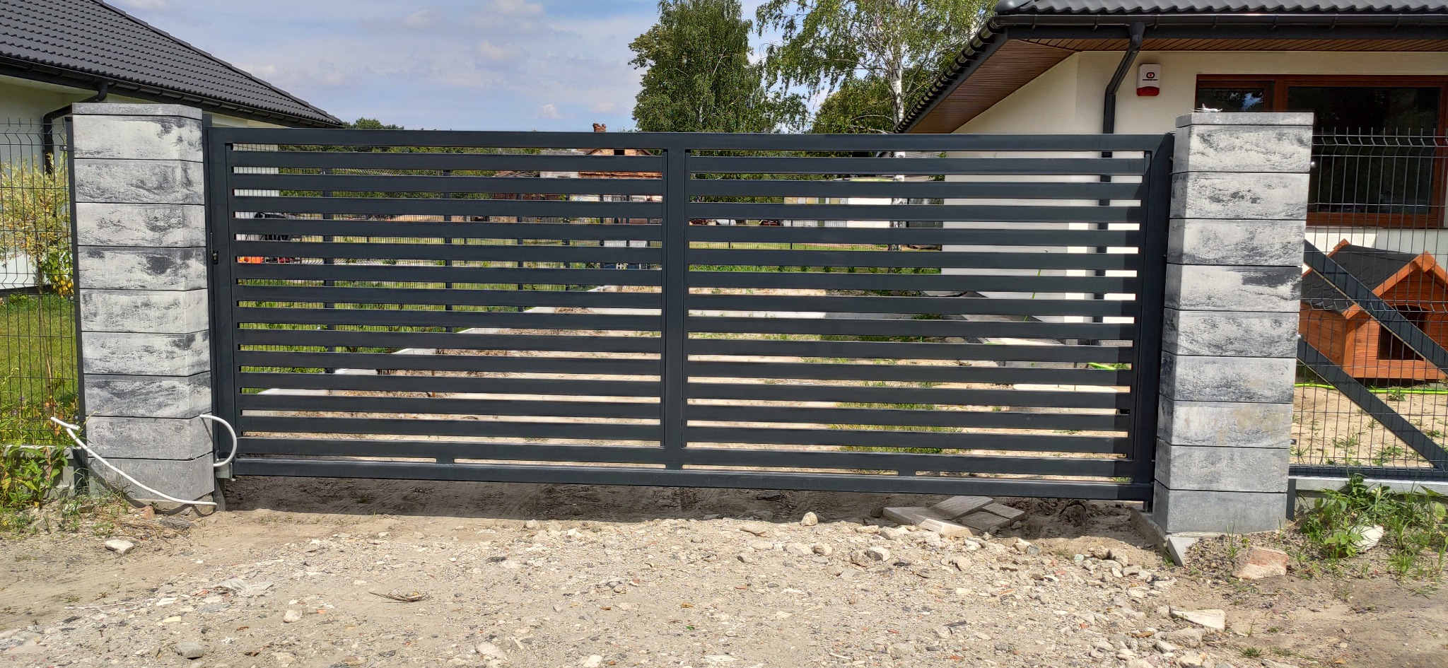 Nowoczesna, przesuwna brama z poziomych, szarych profili, osadzona na słupkach z szarej kostki betonowej, widoczny fragment posesji i sąsiedniego domu.