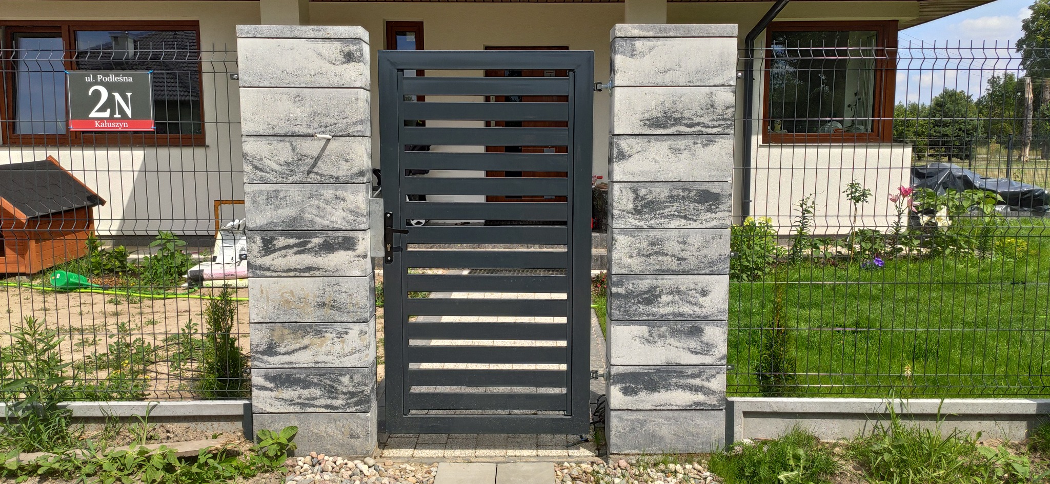 Nowoczesna, szara brama z poziomymi szczeblami, osadzona między dwoma słupkami z szarej kostki imitującej kamień, z widocznym adresem na ogrodzeniu z siatki w tle.