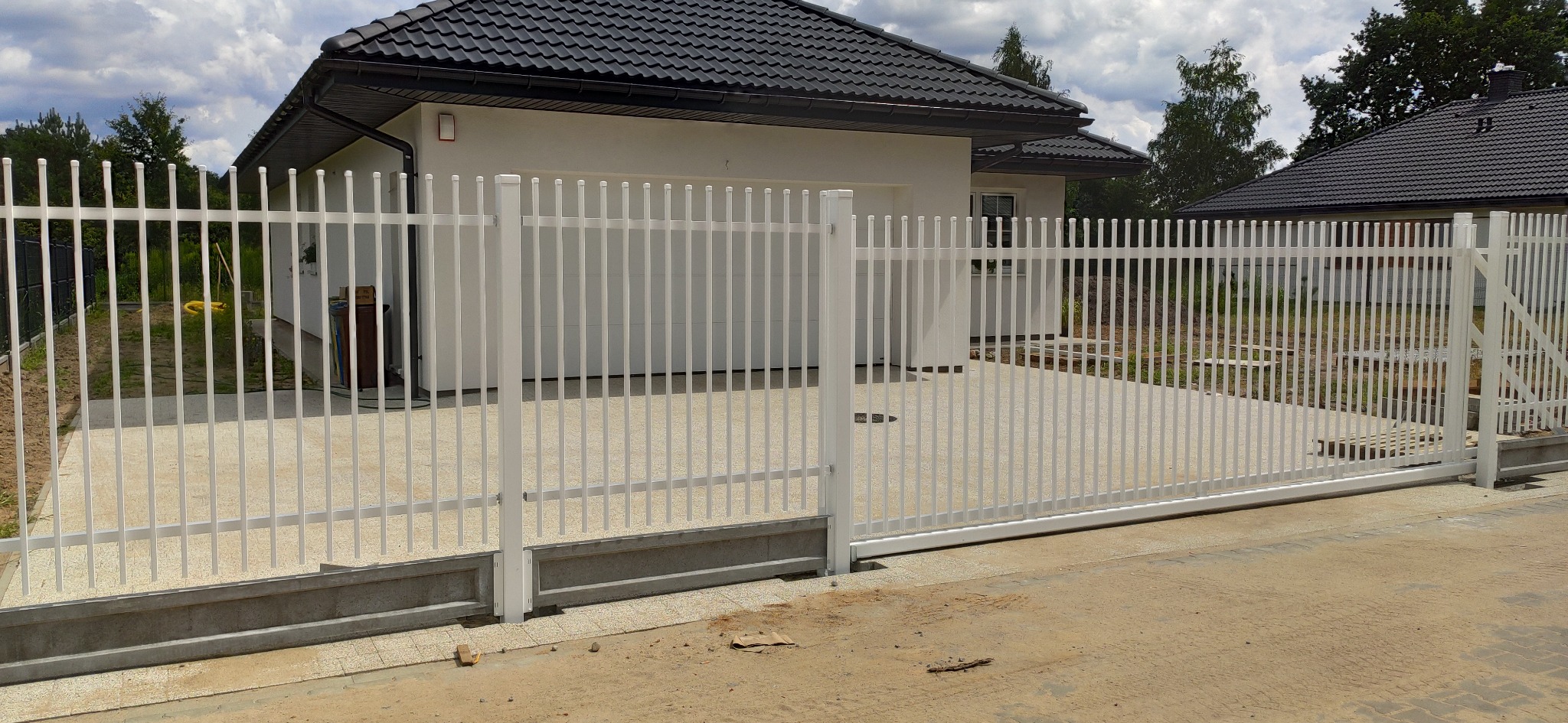 Biała, przesuwna brama panelowa z pionowych prętów na tle jasnego domu z ciemną dachówką.