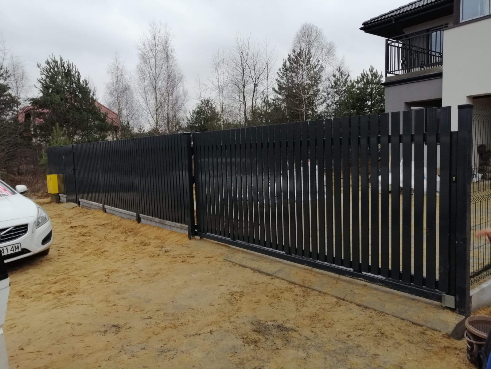 Czarny, pionowy płot panelowy z przesuwną bramą wjazdową, widoczny z boku na piaszczystym podłożu, z fragmentem białego samochodu po lewej stronie i budynkiem w tle.