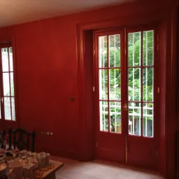 Wnętrze pomalowane na czerwono z widokiem na ogród przez okno balkonowe i okno, na stole zastawa stołowa