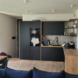 MKDom - Nowoczesna kuchnia z czarnymi szafkami i drewnianym blatem, widok na salon z niebieską sofą, okno balkonowe z widokiem na zieleń, minimalistyczne oświetlenie sufitowe.