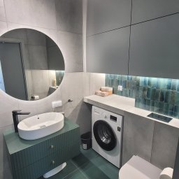 MKDom - Nowoczesna łazienka w odcieniach szarości i zieleni z okrągłym podświetlanym lustrem, umywalką na szafce z ryflowanym frontem, pralką pod blatem i mozaiką nad nią.