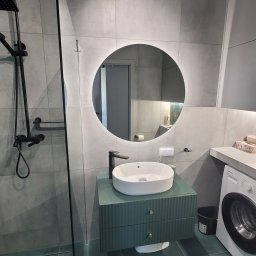 MKDom - Nowoczesna łazienka w odcieniach szarości i zieleni z okrągłym lustrem z podświetleniem LED, minimalistyczną umywalką na szafce z frezowanymi frontami i czarną armaturą, obok pralka.