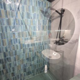 MKDom - Nowoczesna kabina prysznicowa z turkusowymi, pionowo ułożonymi płytkami, czarną armaturą i szklaną ścianką, widoczna biała umywalka i butelka kosmetyku.