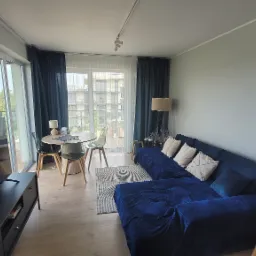 Jasne wnętrze salonu z niebieską sofą narożną, okrągłym stołem z transparentnymi krzesłami i dużym oknem balkonowym z ciemnymi zasłonami.