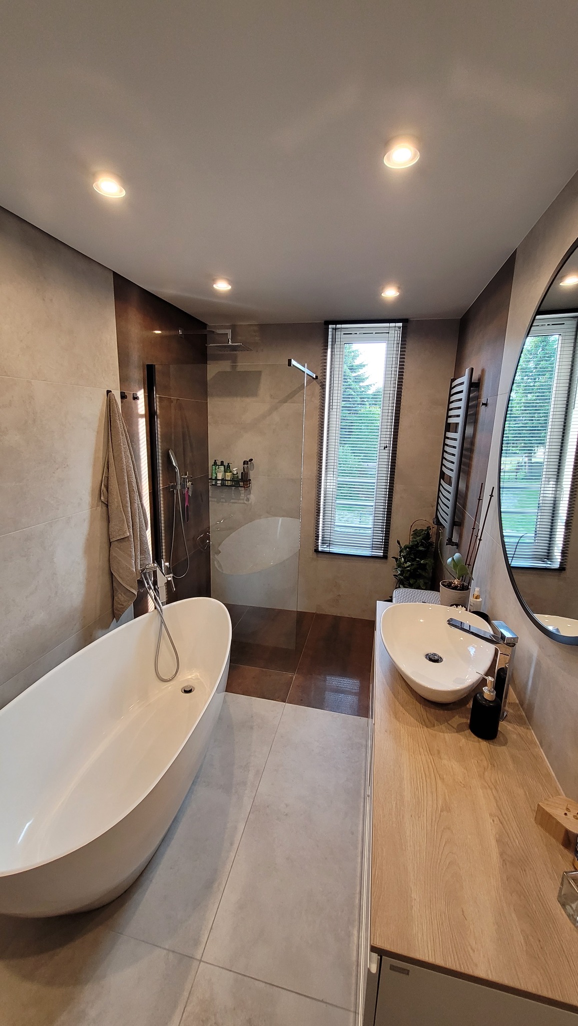 Nowoczesna łazienka z wolnostojącą wanną, kabiną prysznicową typu walk-in, umywalką nablatową na drewnianym blacie i dużym okrągłym lustrem, oświetlona punktowymi lampami sufitowymi.