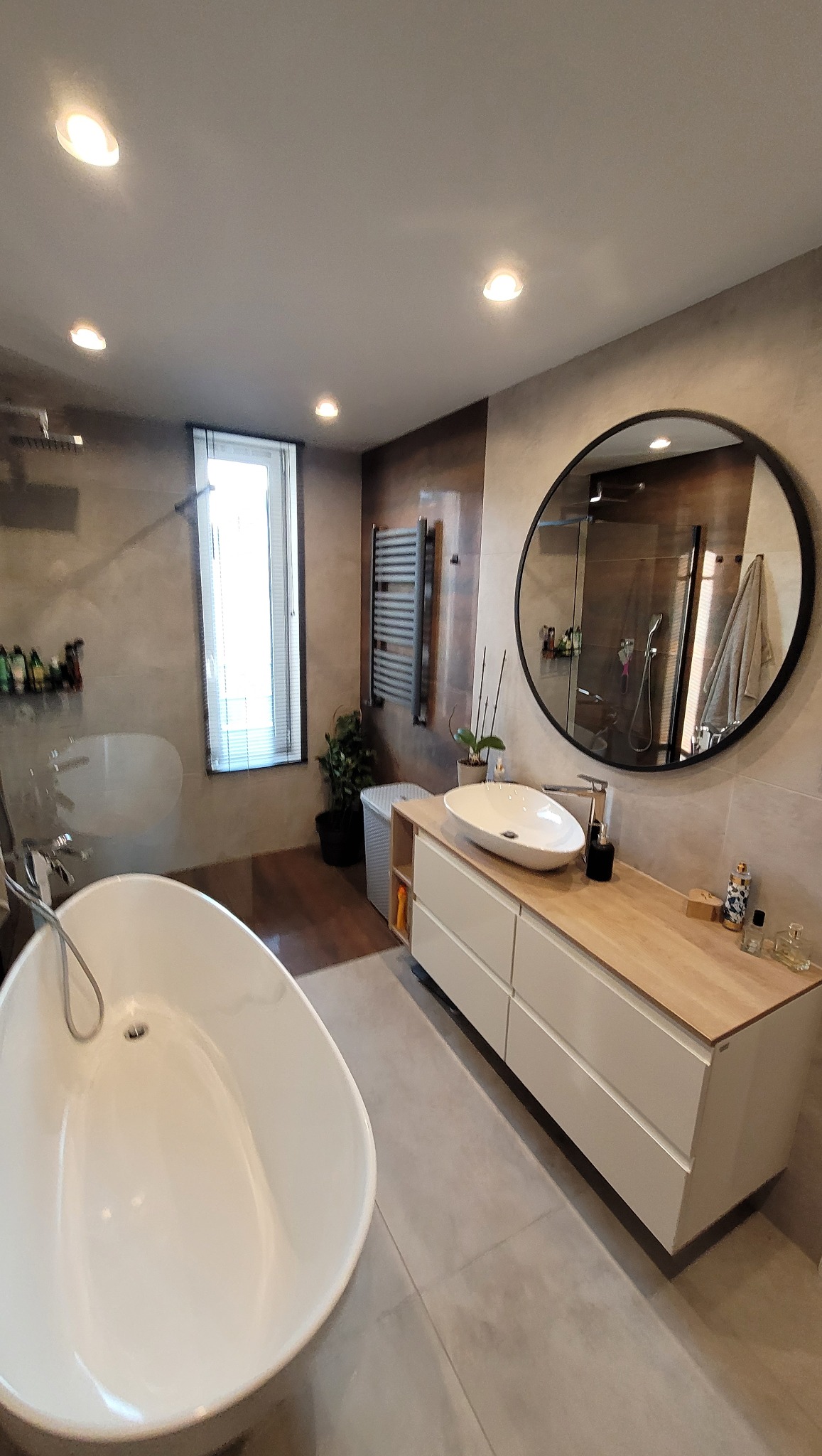 Nowoczesna łazienka z wolnostojącą wanną, umywalką nablatową na szafce z drewnianym blatem, okrągłym lustrem i grzejnikiem drabinkowym na ścianie z efektem betonu.