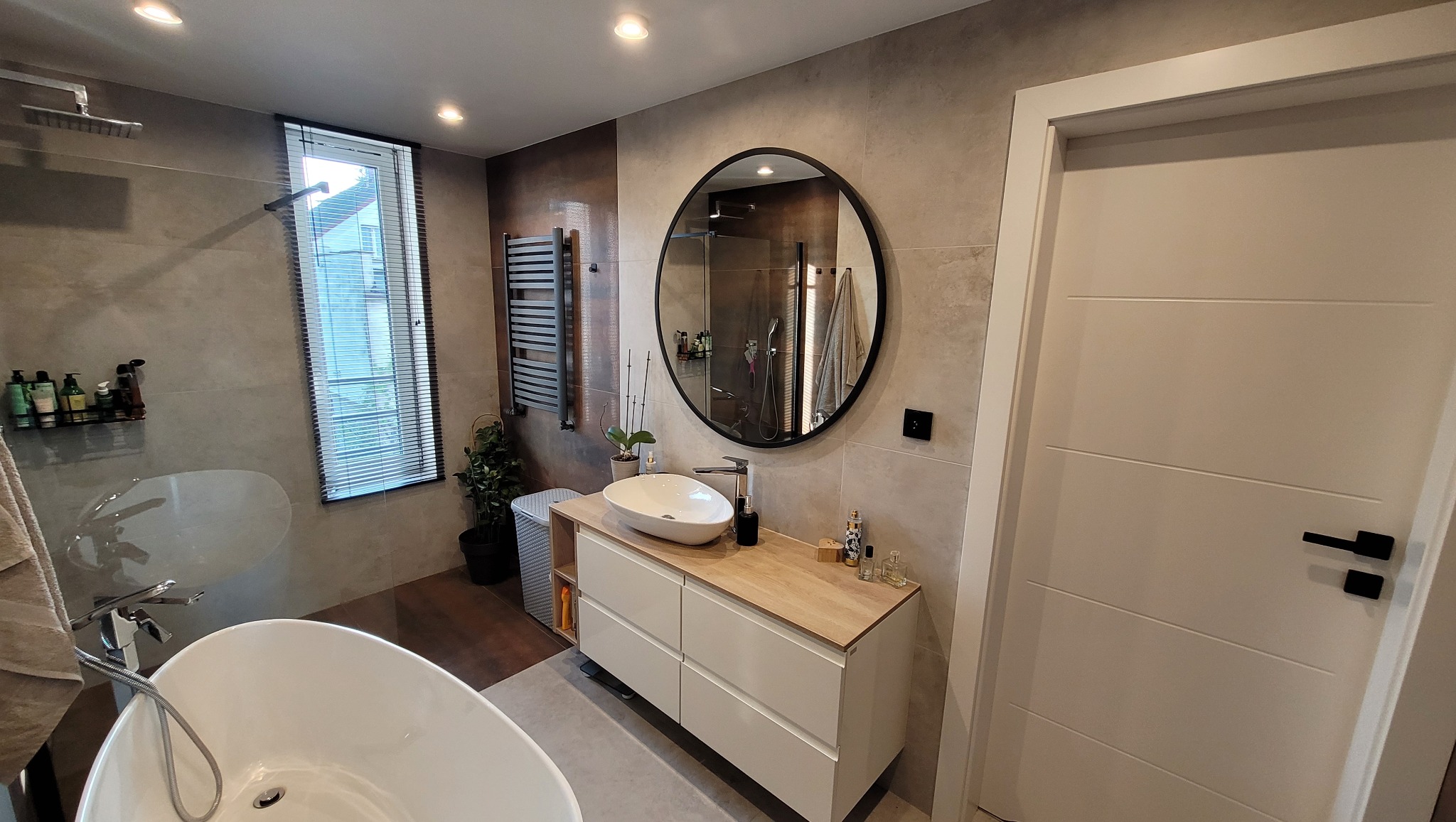 Nowoczesna łazienka z wolnostojącą wanną, umywalką nablatową na szafce z drewnianym blatem, okrągłym lustrem w czarnej ramie i grzejnikiem drabinkowym na ścianie z szarych płytek.