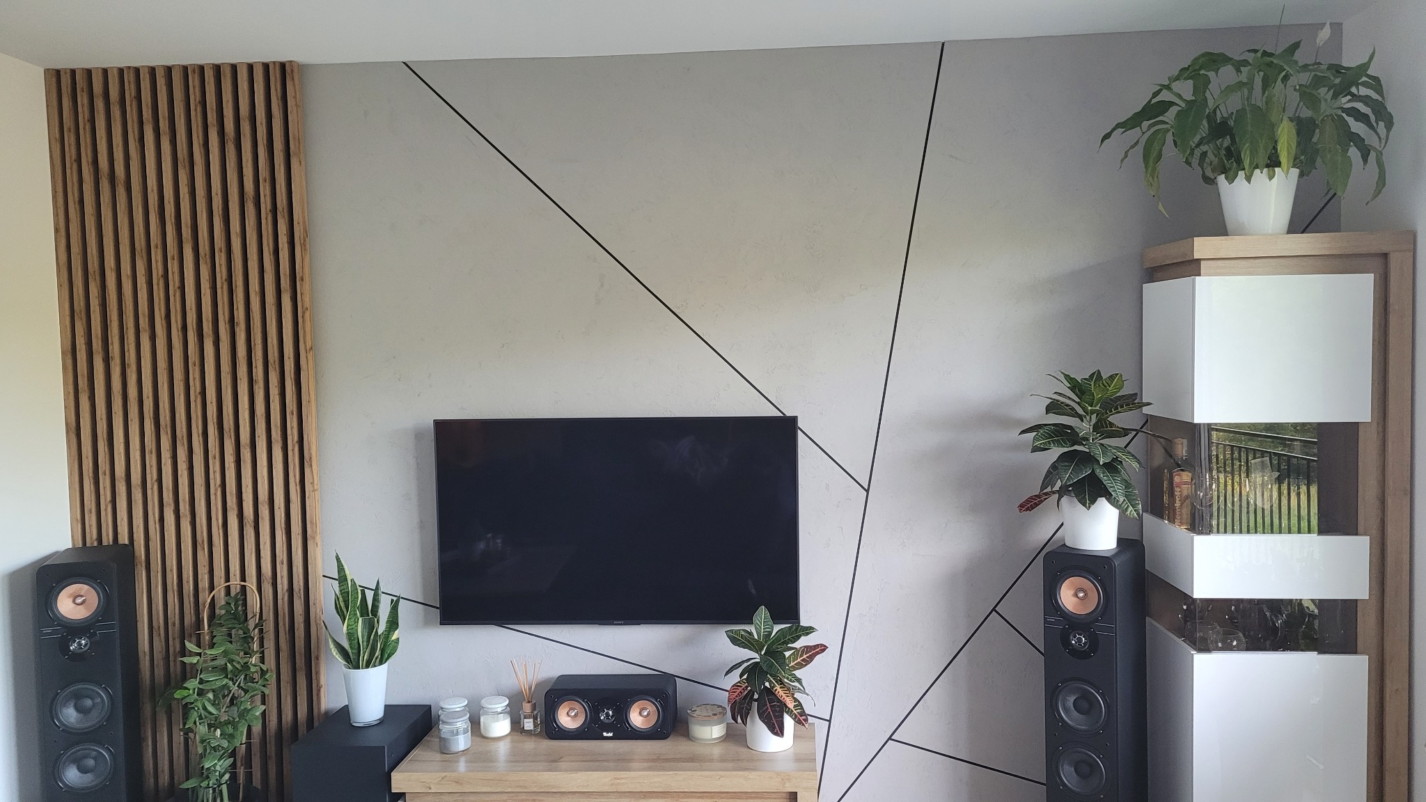 Szara ściana z geometrycznym wzorem z czarnych linii, drewniana lamelowa ściana, telewizor, głośniki i rośliny doniczkowe w nowoczesnym salonie.