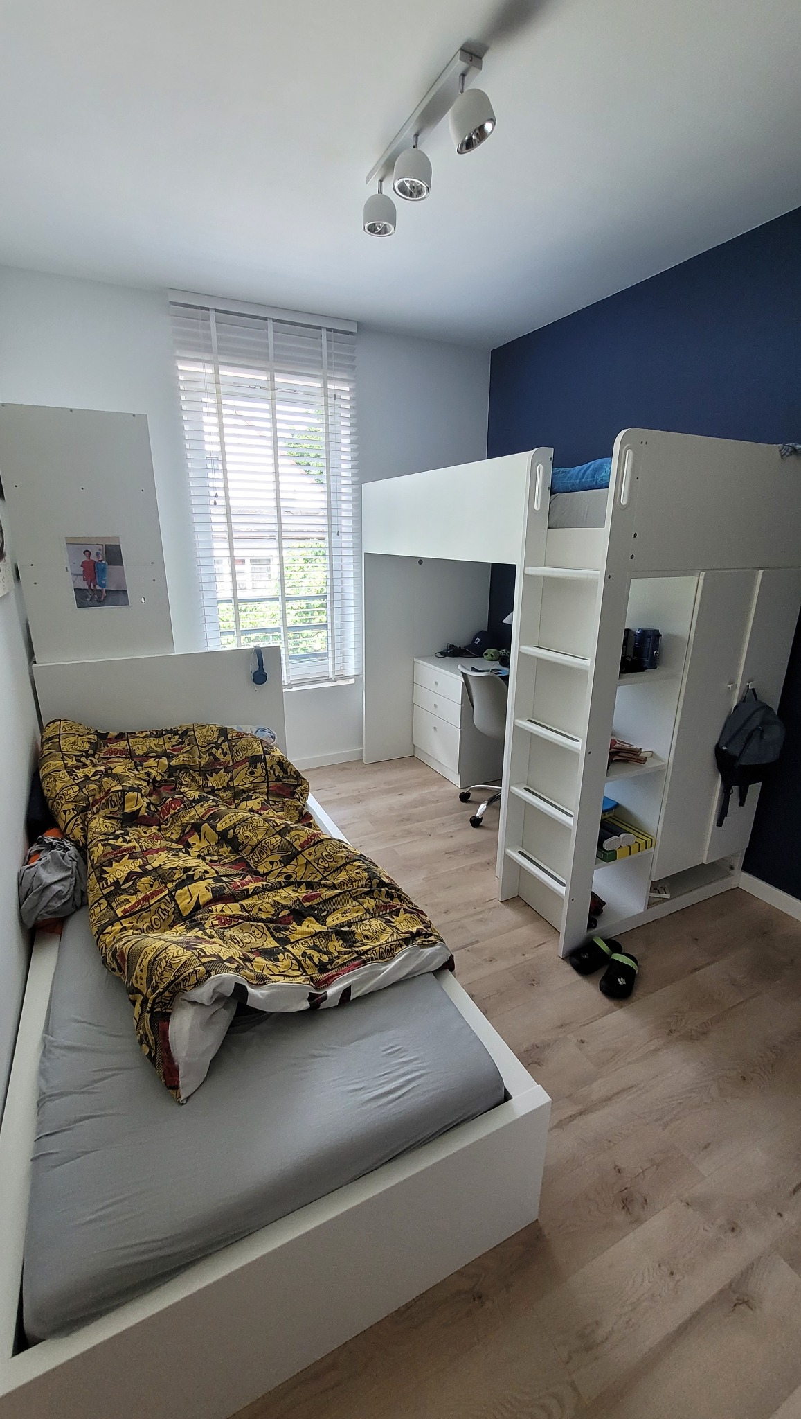Pokój dziecięcy z białym łóżkiem piętrowym z biurkiem, pojedynczym łóżkiem z szarą pościelą i kolorową kołdrą w komiksowe wzory, drewniana podłoga i granatowa ściana.