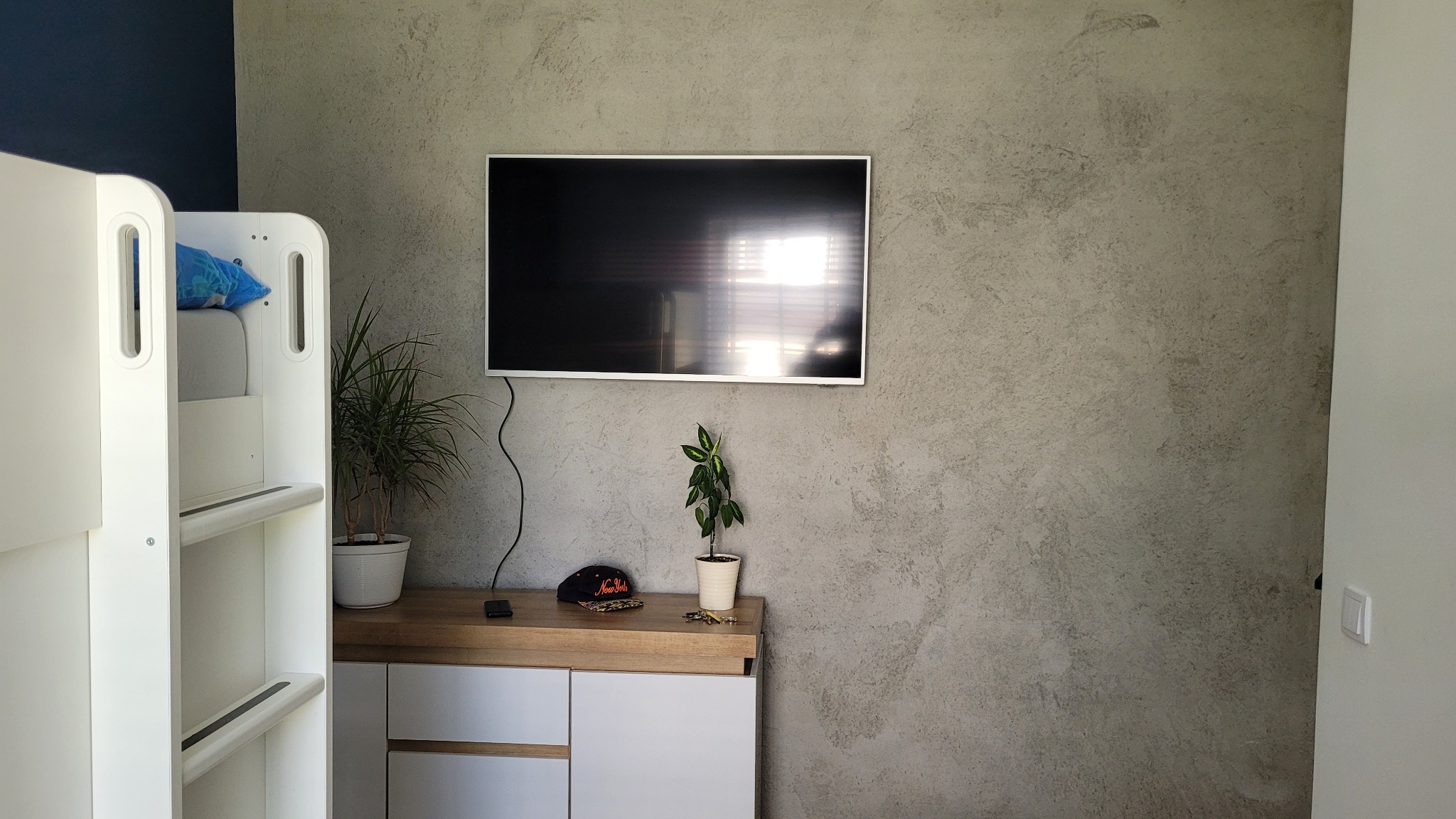 Pokój z łóżkiem piętrowym, telewizorem na ścianie pokrytej dekoracyjną szarą fakturą, szafką z roślinami i czapką.