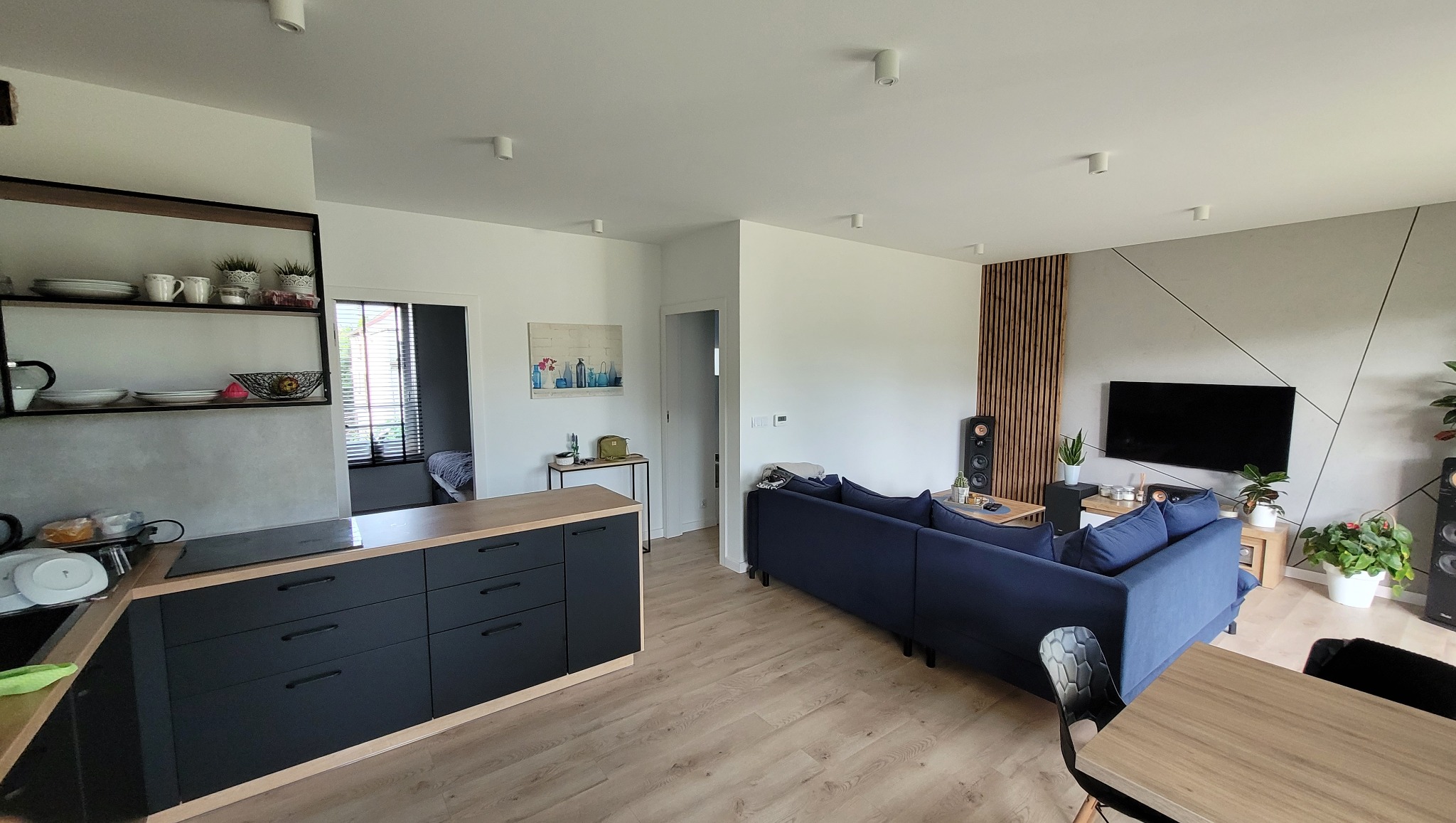 Przestronne, jasne wnętrze po remoncie w Rybniku: otwarta kuchnia z czarnymi szafkami i drewnianym blatem, połączona z salonem z dużą, granatową sofą i telewizorem na ścianie z geometrycznym wzorem.