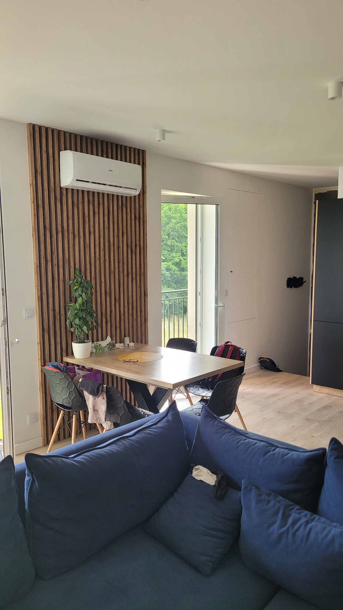 Nowoczesne wnętrze: drewniana ściana z lamelami, klimatyzator, stół z krzesłami i widok na balkon przez przeszklone drzwi, fragment niebieskiej sofy na pierwszym planie.