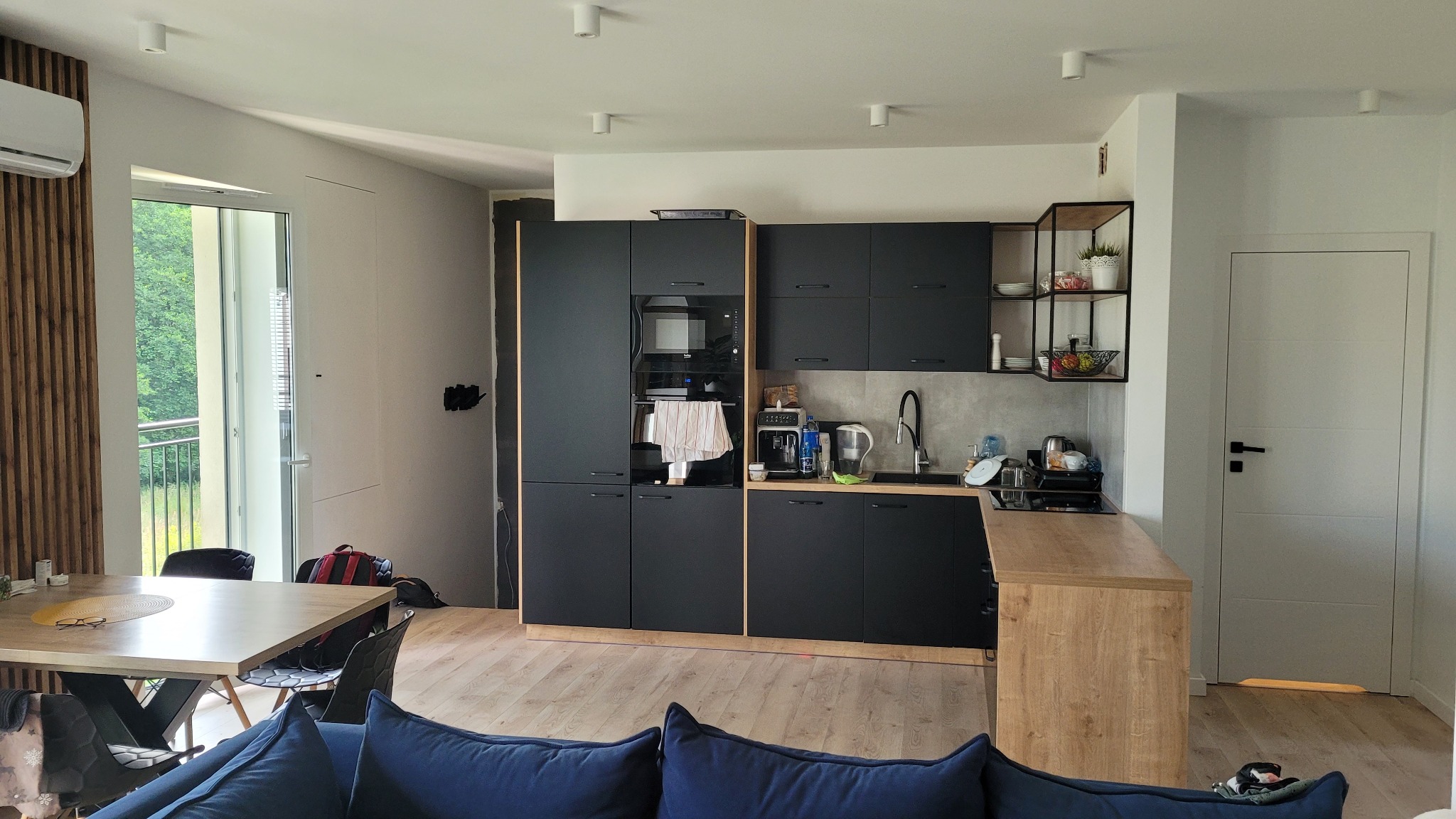 Nowoczesna kuchnia z czarnymi szafkami i drewnianym blatem, widok na salon z niebieską sofą, okno balkonowe z widokiem na zieleń, minimalistyczne oświetlenie sufitowe.