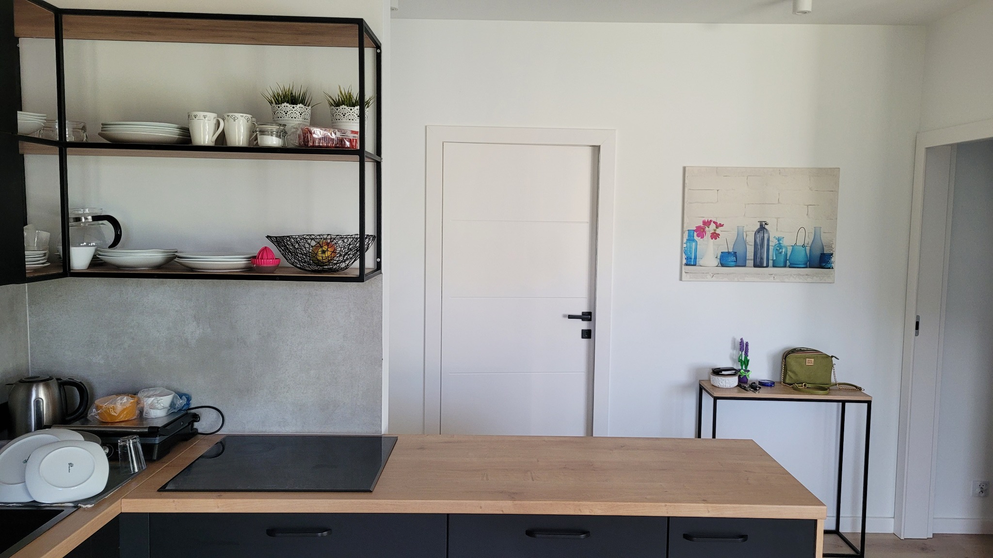 Jasna kuchnia z drewnianym blatem, czarnymi szafkami, półkami w metalowej ramie i białymi ścianami. Na półkach naczynia i dekoracje. Na ścianie obraz z niebieskimi wazonami, a obok stolik z torebką...