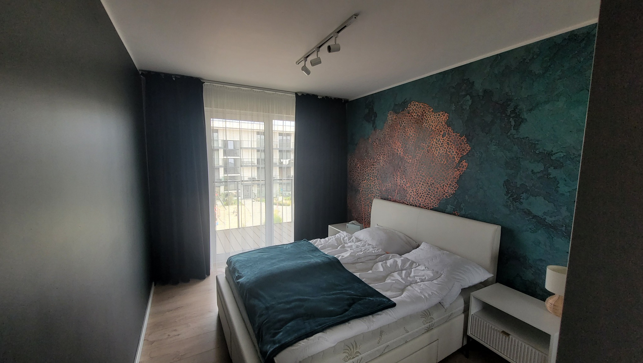 Sypialnia z łóżkiem kontynentalnym, ciemną ścianą i ścianą akcentową z wzorem koralowca, oświetleniem punktowym i wyjściem na balkon.