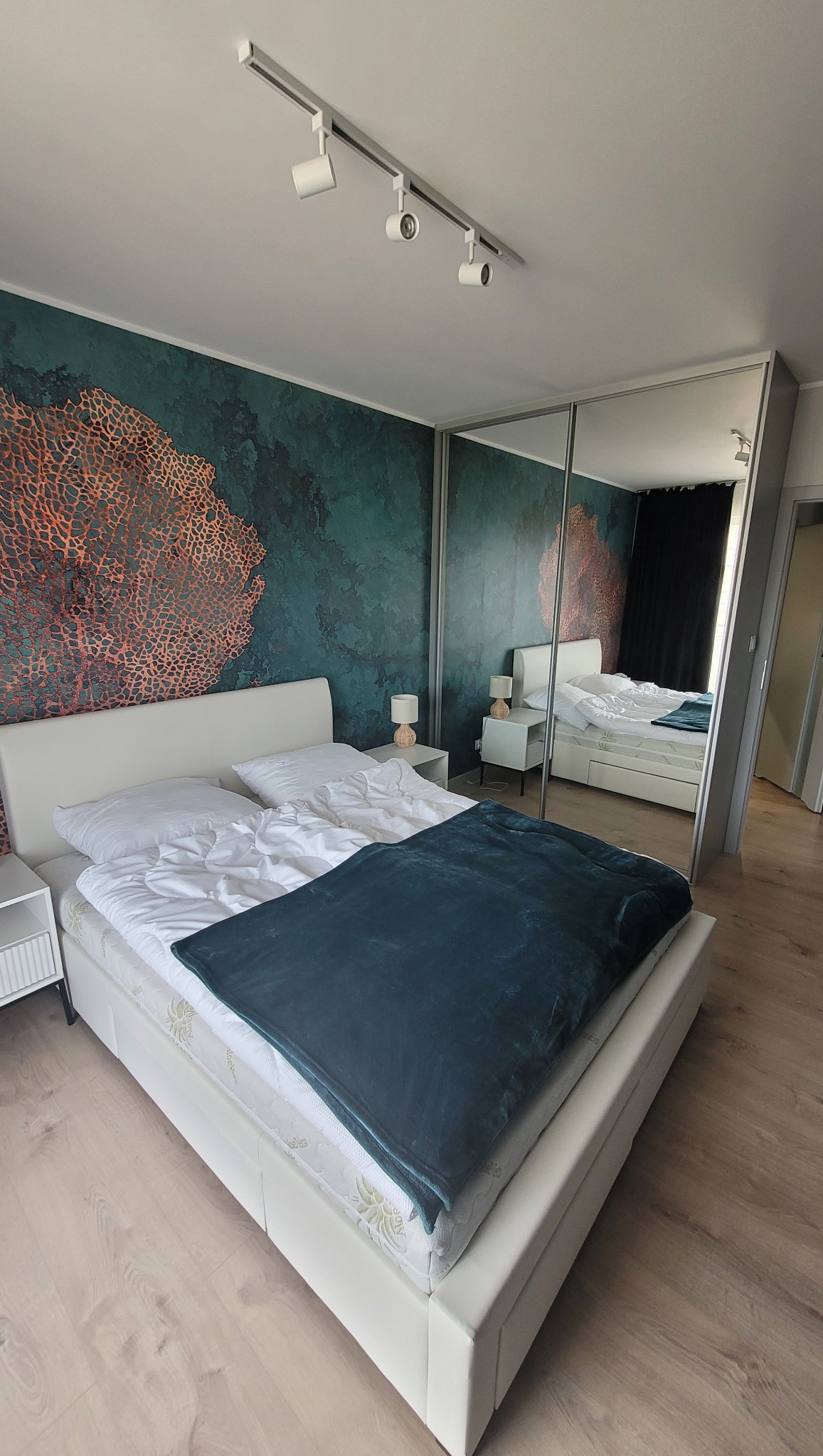 Sypialnia z białym łóżkiem, zieloną ścianą z wzorem koralowca i lustrzaną szafą przesuwną, odbijającą drugie łóżko w pokoju.