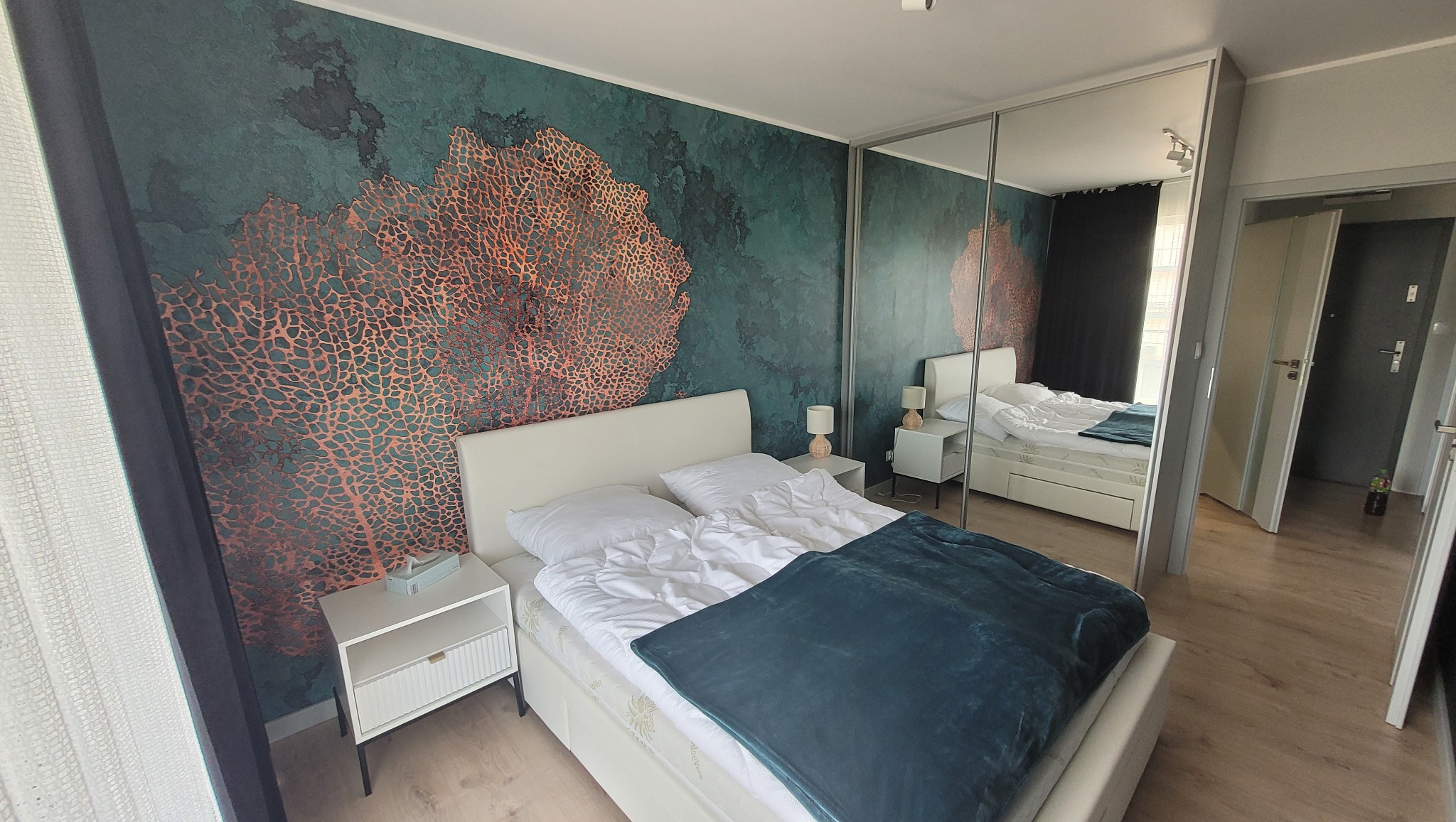 Sypialnia z łóżkiem, szafkami nocnymi i dekoracyjną ścianą w odcieniach zieleni i miedzi, wzór przypominający koralowiec, odbicie pokoju w lustrzanych drzwiach szafy w tle.