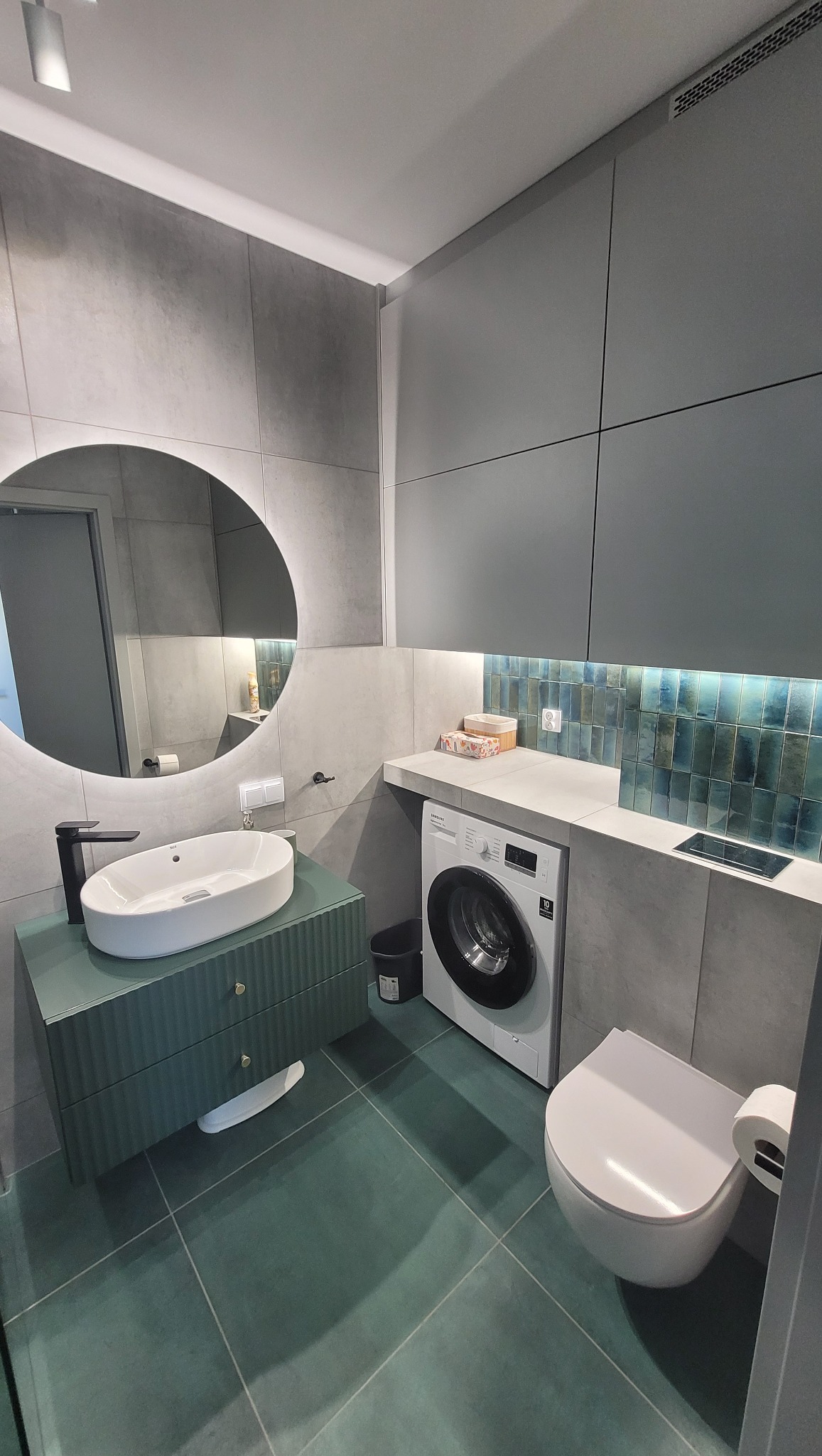 Nowoczesna łazienka w odcieniach szarości i zieleni z okrągłym podświetlanym lustrem, umywalką na szafce z ryflowanym frontem, pralką pod blatem i mozaiką nad nią.
