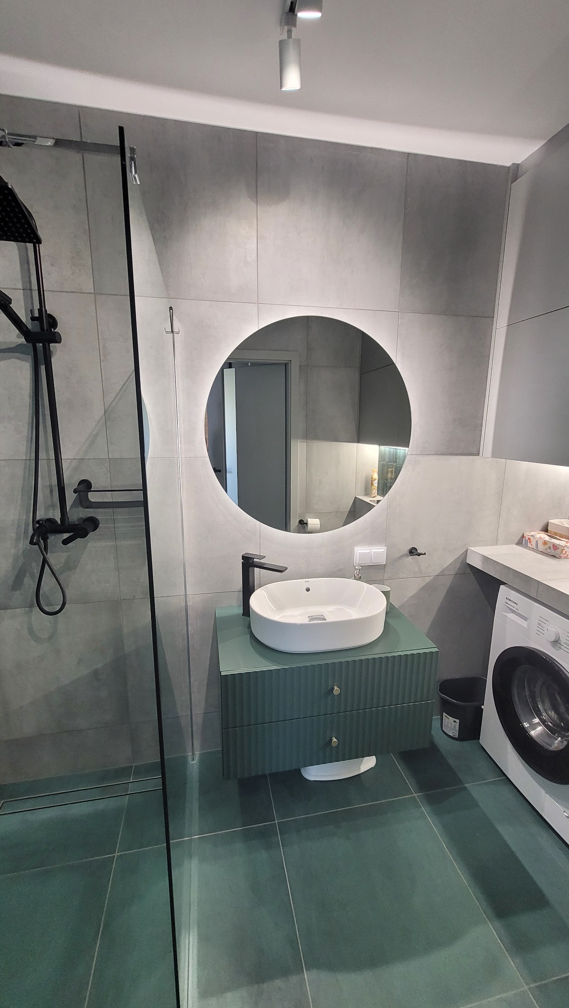 Nowoczesna łazienka w odcieniach szarości i zieleni z okrągłym lustrem z podświetleniem LED, minimalistyczną umywalką na szafce z frezowanymi frontami i czarną armaturą, obok pralka.