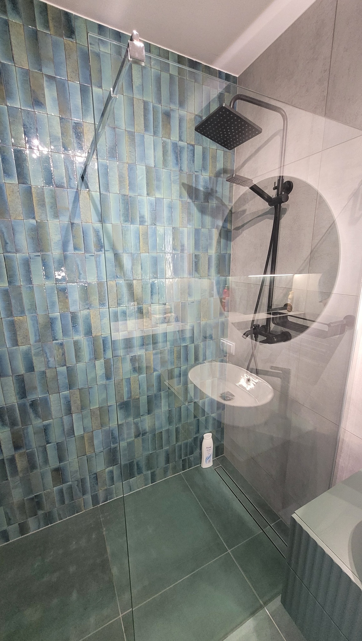 Nowoczesna kabina prysznicowa z turkusowymi, pionowo ułożonymi płytkami, czarną armaturą i szklaną ścianką, widoczna biała umywalka i butelka kosmetyku.