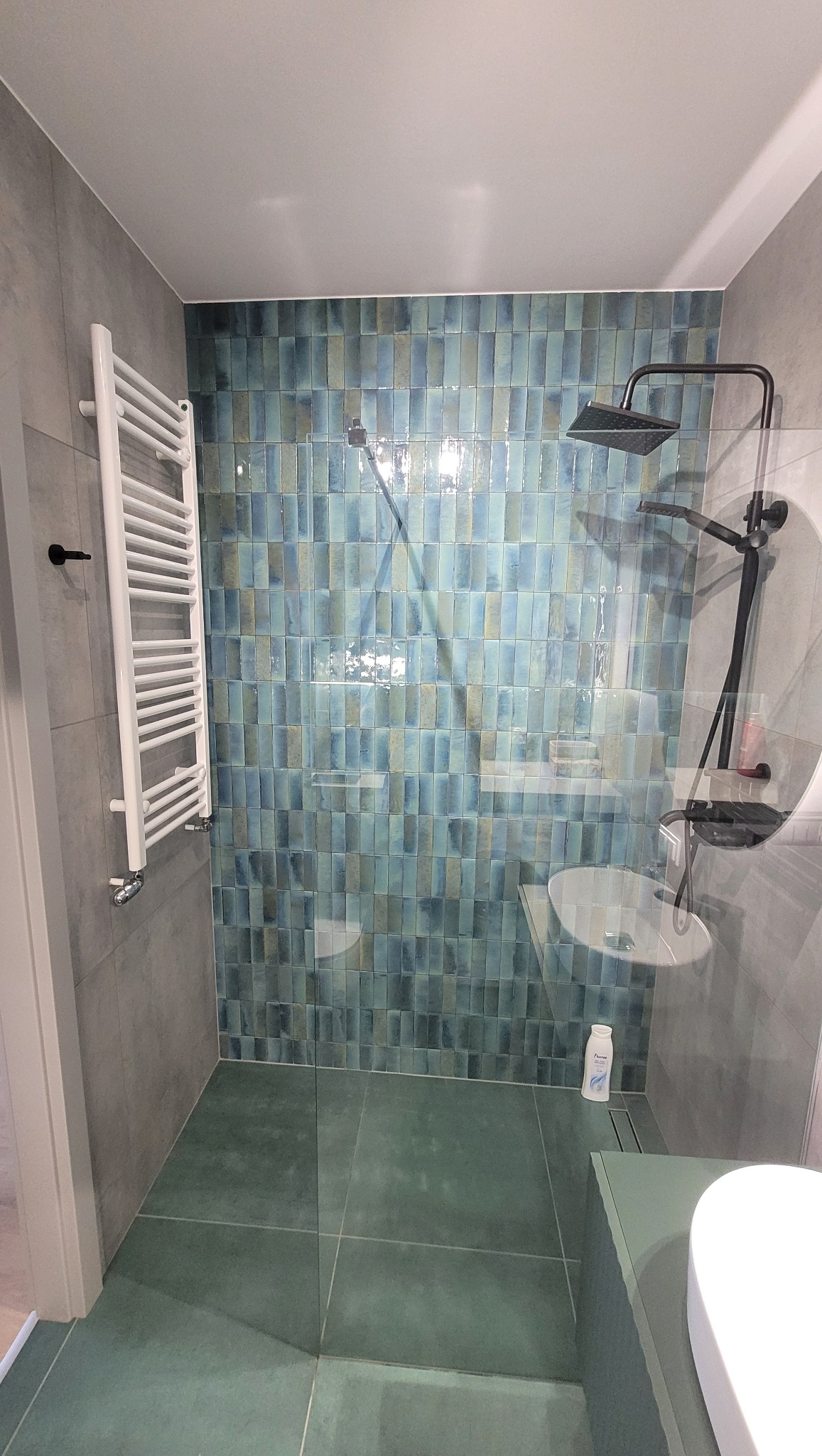 Nowoczesna łazienka z kabiną prysznicową typu walk-in, wykończona turkusową mozaiką na ścianie, szarymi płytkami na pozostałych ścianach i podłodze, z białym grzejnikiem drabinkowym i czarną...